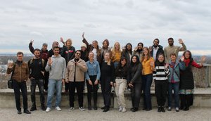 The photo shows a group of people standing in two rows at the edge of a viewing platform, waving at the camera. The back row is standing on the raised railing. In the background, a cityscape with a cloudy sky can be seen.