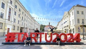 Photo of a paved square in the city of Tartu, surrounded by buildings. In the foreground a sculpture of red letters: #HARTU2024. Two students are sitting in front of the sculpture. One student is sitting on the letter U and has her arms outstretched. Another student is standing behind the sculpture between the letters R and T.