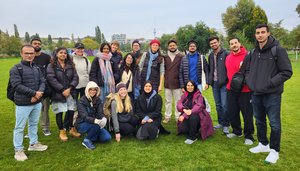 A close-up photo of a group of students and faculty members on a lawn. One row is kneeling, one row is standing, and all are facing the camera. Trees and buildings can be seen in the background