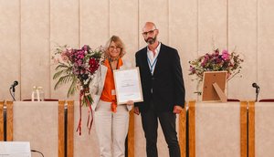 Photo. Prof. Ingrid Schegk can be seen in the center of the picture, holding a certificate and a bouquet of flowers. Frederico Meireles is standing to her right. A long table and a beige wall can be seen in the background.
