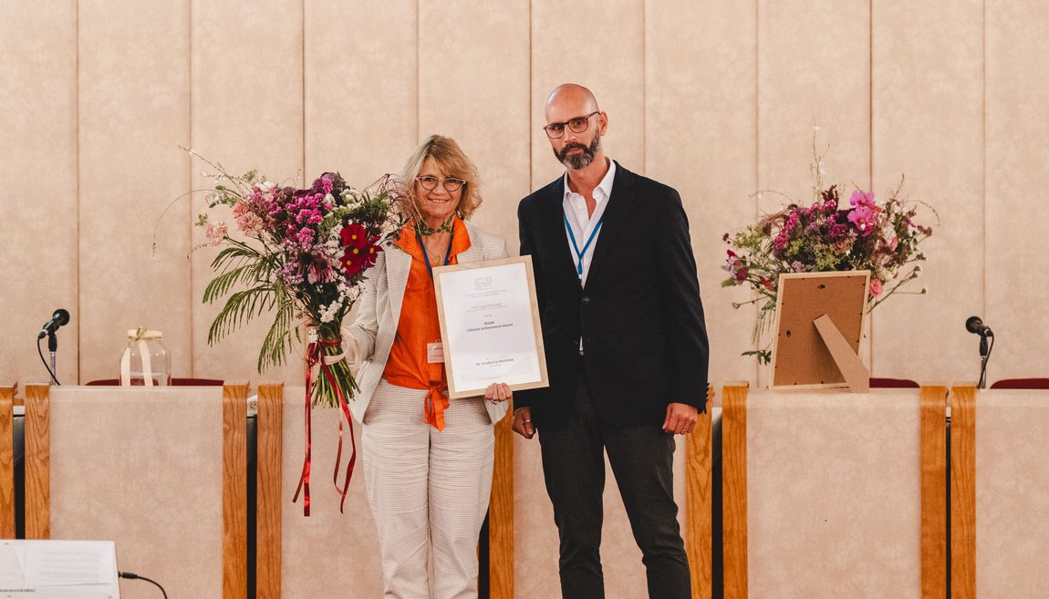 Photo. Prof. Ingrid Schegk can be seen in the center of the picture, holding a certificate and a bouquet of flowers. Frederico Meireles is standing to her right. A long table and a beige wall can be seen in the background.
