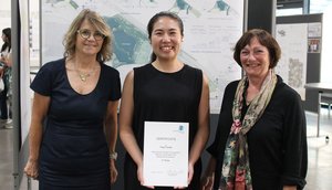 Photo of Yuga Tanaka with her supervisors Prof. Ingrid Schegk (left) and Prof. Cornelia Bott (right) in the foreground. In the background a movable wall with a plan.