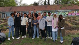 Photo - A group of students stand on a meadow and look into the camera. In the background is a garden and a building with graffiti on the wall.