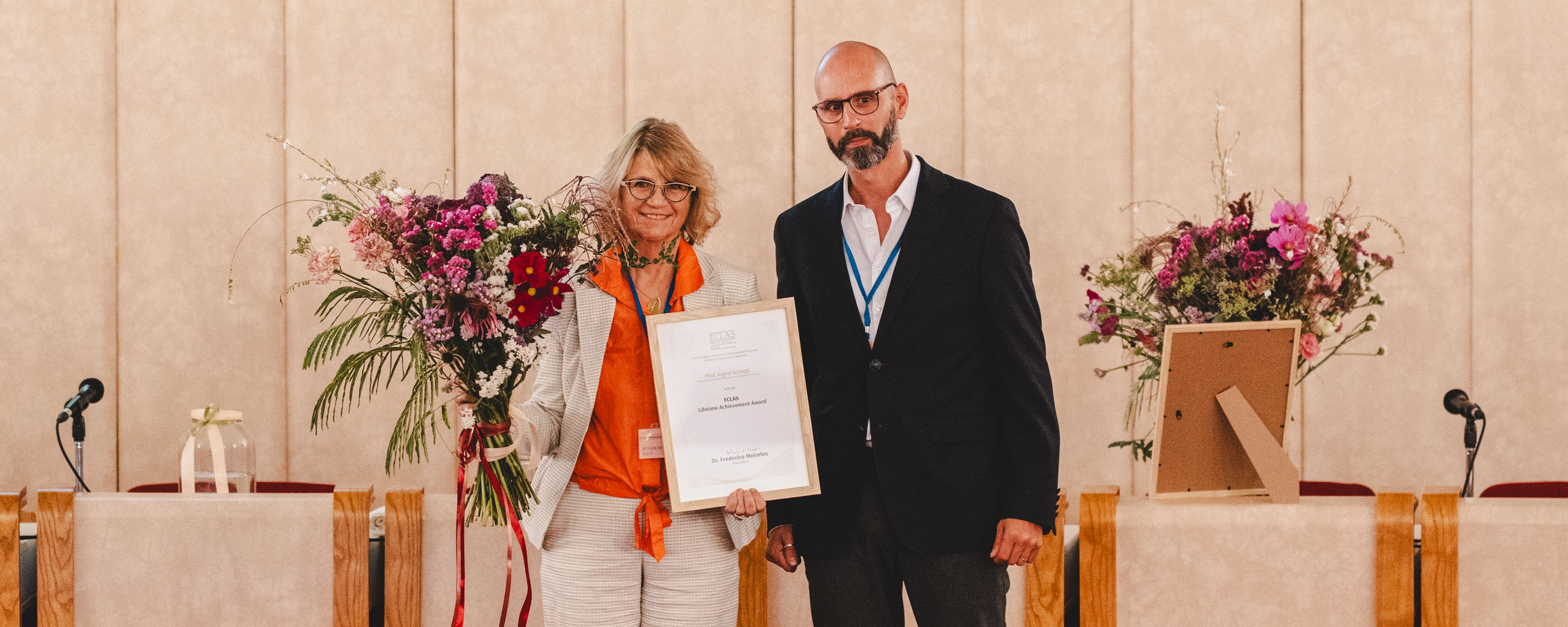 slide - Photo. Prof. Ingrid Schegk can be seen in the center of the picture, holding a certificate and a bouquet of flowers. Frederico Meireles is standing to her right. A long table and a beige wall can be seen in the background.