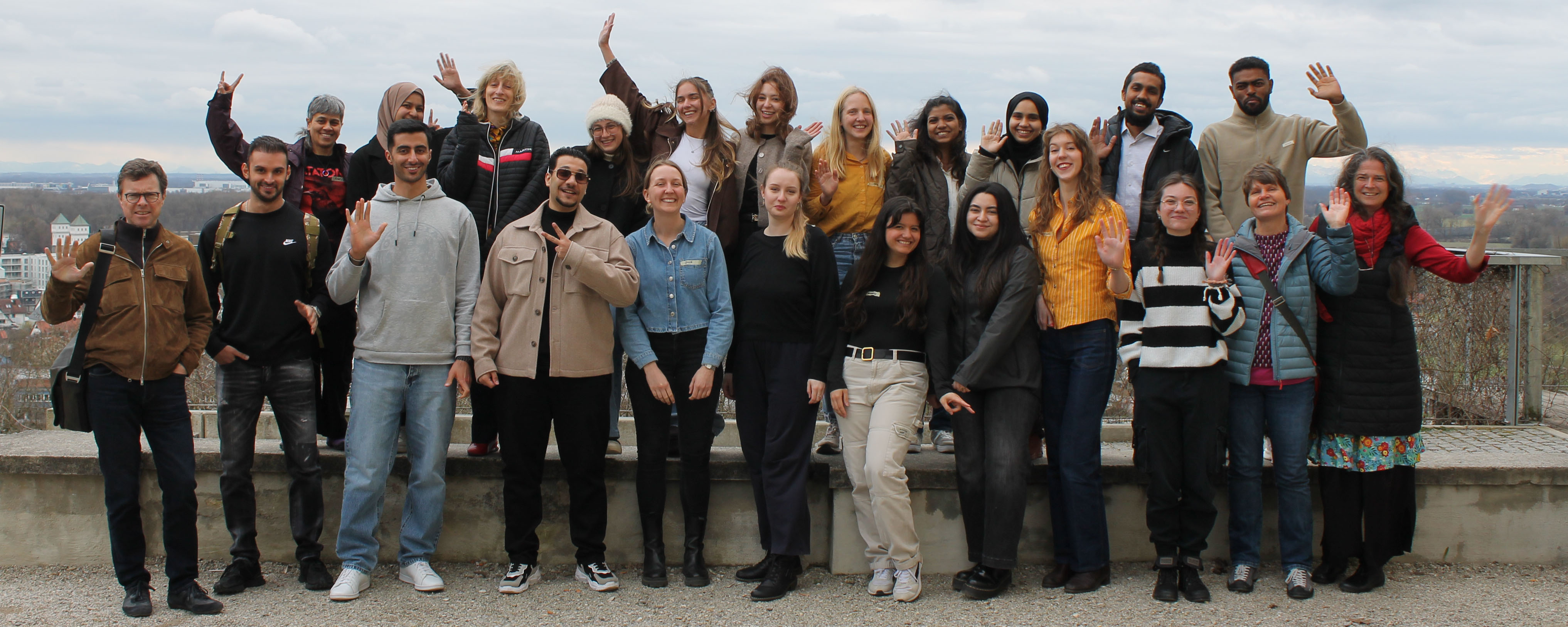 slide - The photo shows a group of people standing in two rows at the edge of a viewing platform, waving at the camera. The back row is standing on the raised railing. In the background, a cityscape with a cloudy sky can be seen.