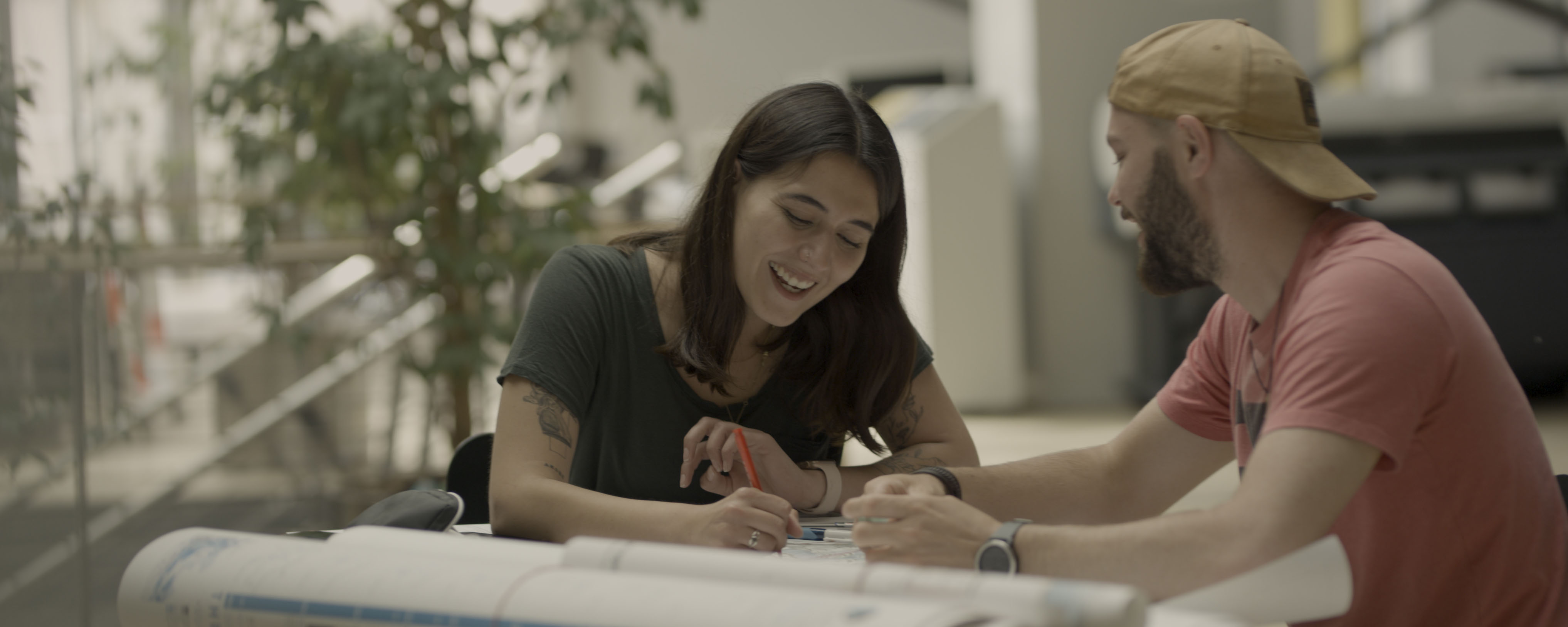 slide - The photo shows two students sitting at a table talking to each other. There are plans on the table. The room furnishings are blurred in the background.