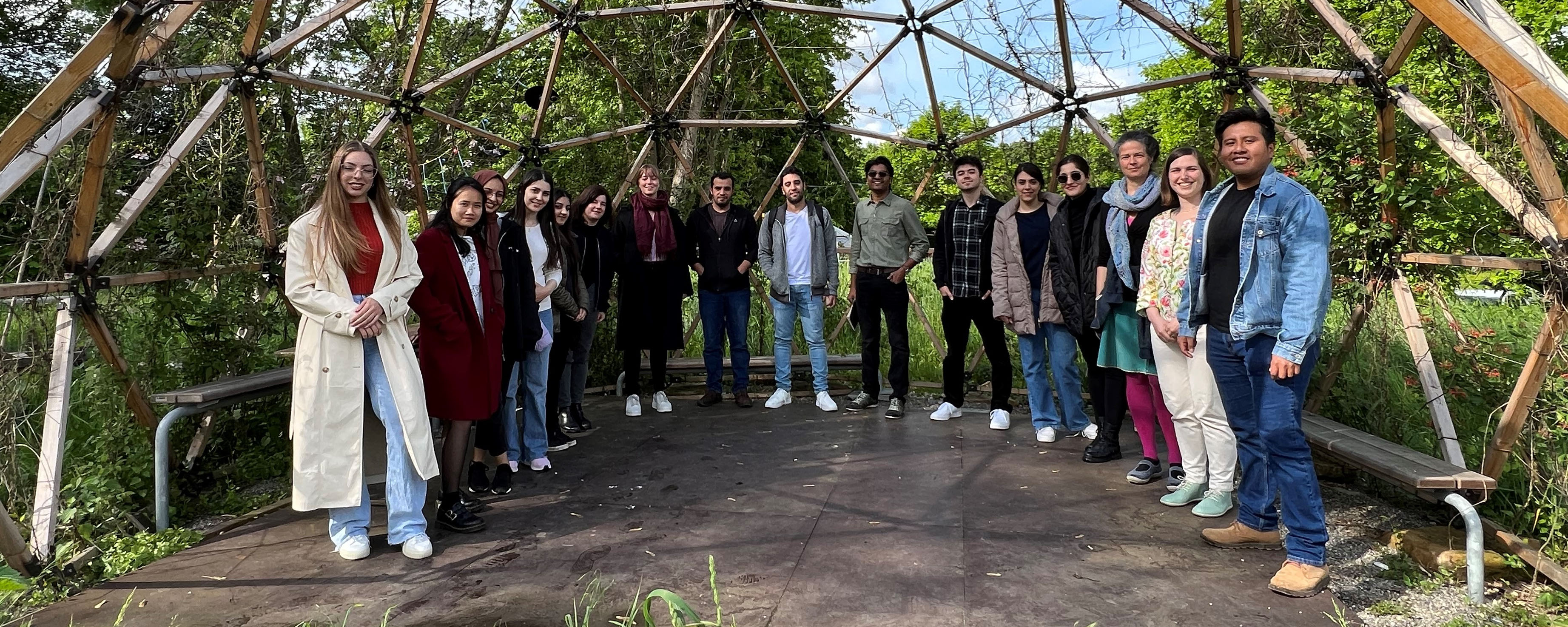 slide - photo group of students around a slabbed square, square on a meadow, covered by a wooden dome, trees in the background.