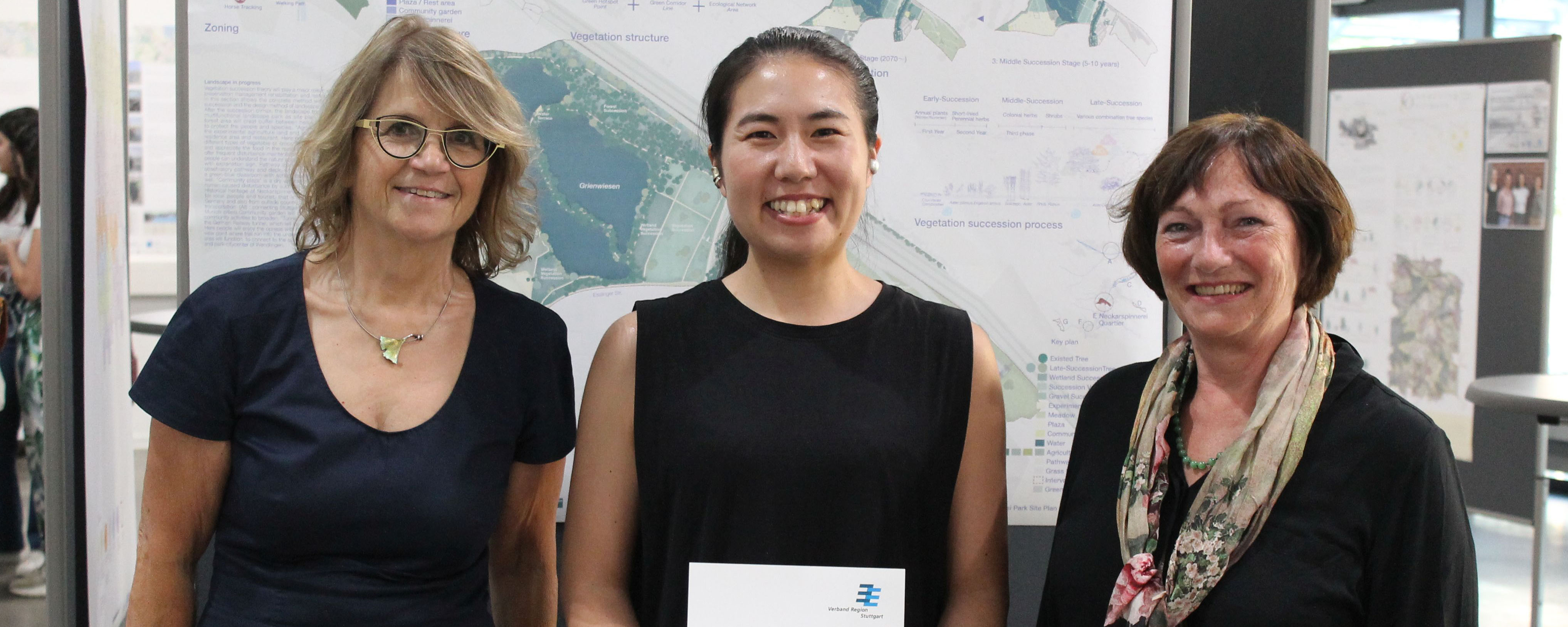 slide - photo of Yuga Tanaka with her supervisors Prof. Ingrid Schegk (left) and Prof. Cornelia Bott (right) in the foreground. In the background a movable wall with a plan.