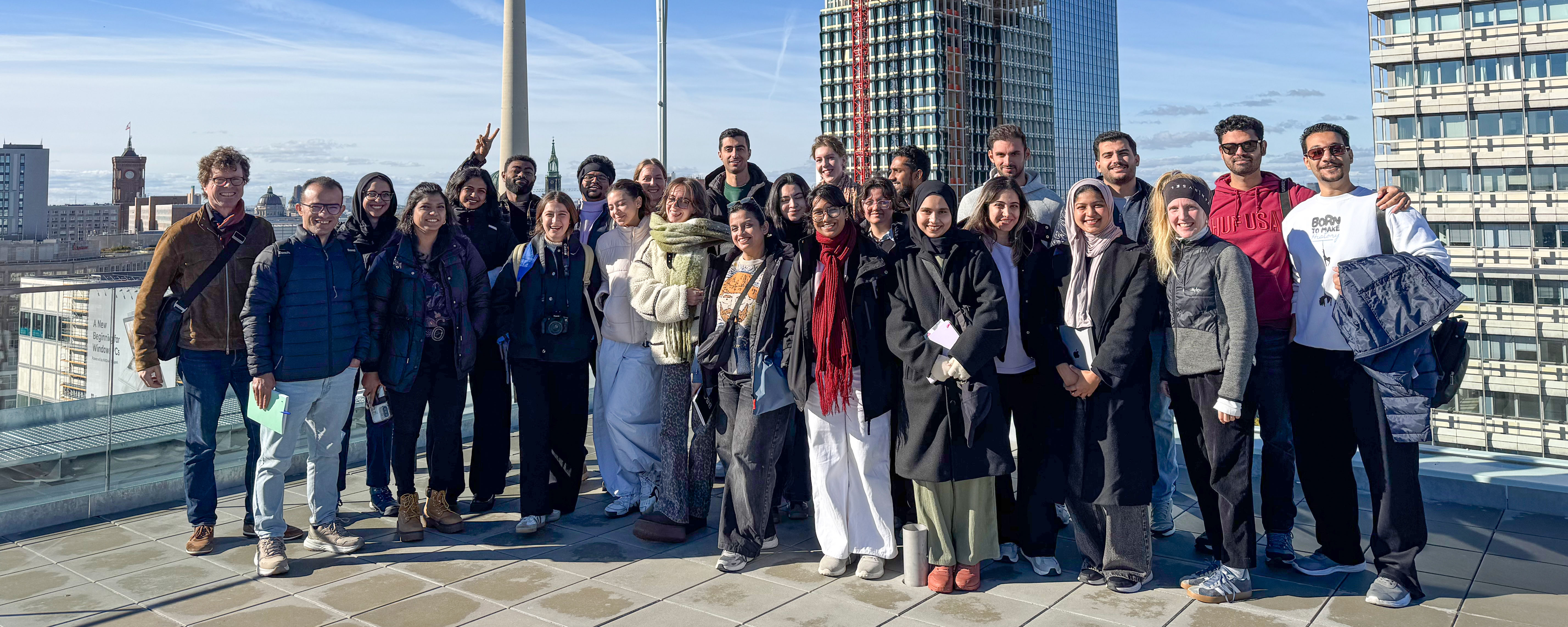 slide - A close-up photo of a group of students and faculty members standing in two rows on a roof terrace. Everyone is facing the camera. Buildings and the sky are visible in the background
