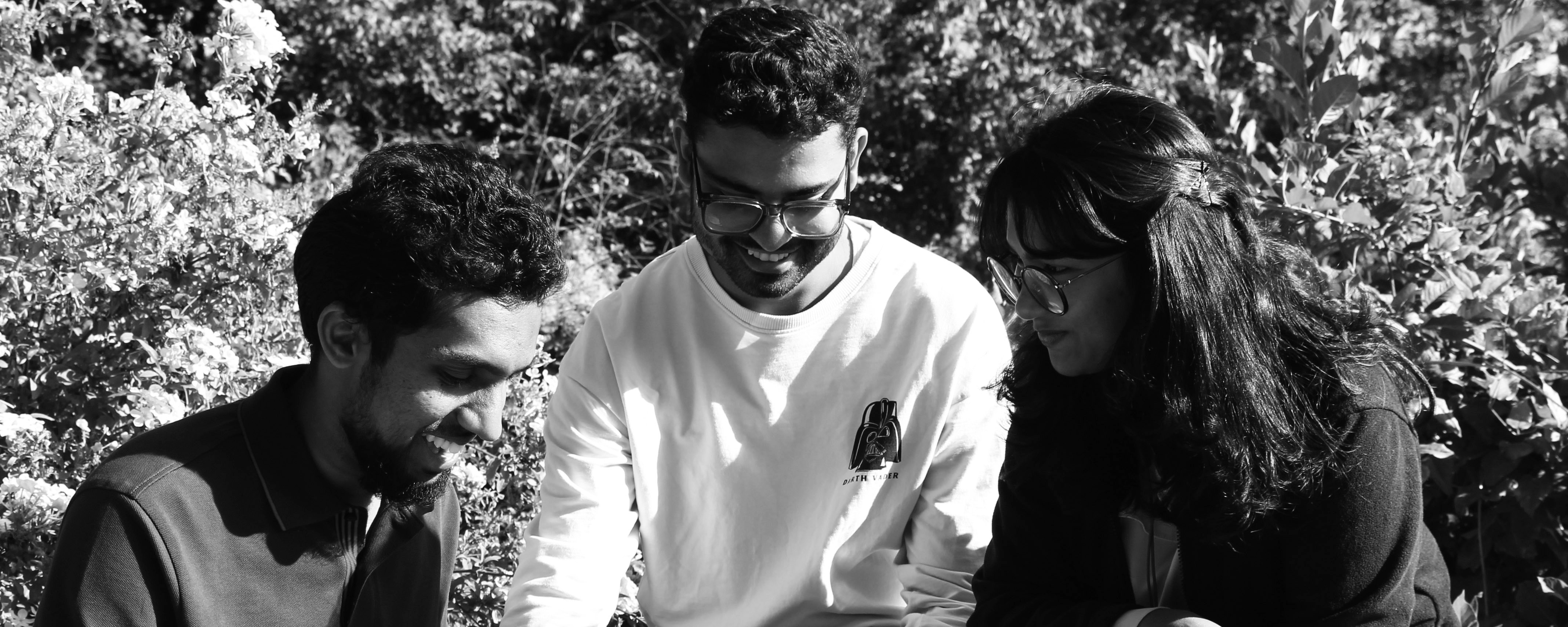 slide - Photo detail in shades of gray: Three IMLA students are sitting next to each other in a park. Shrubs can be seen in the background.