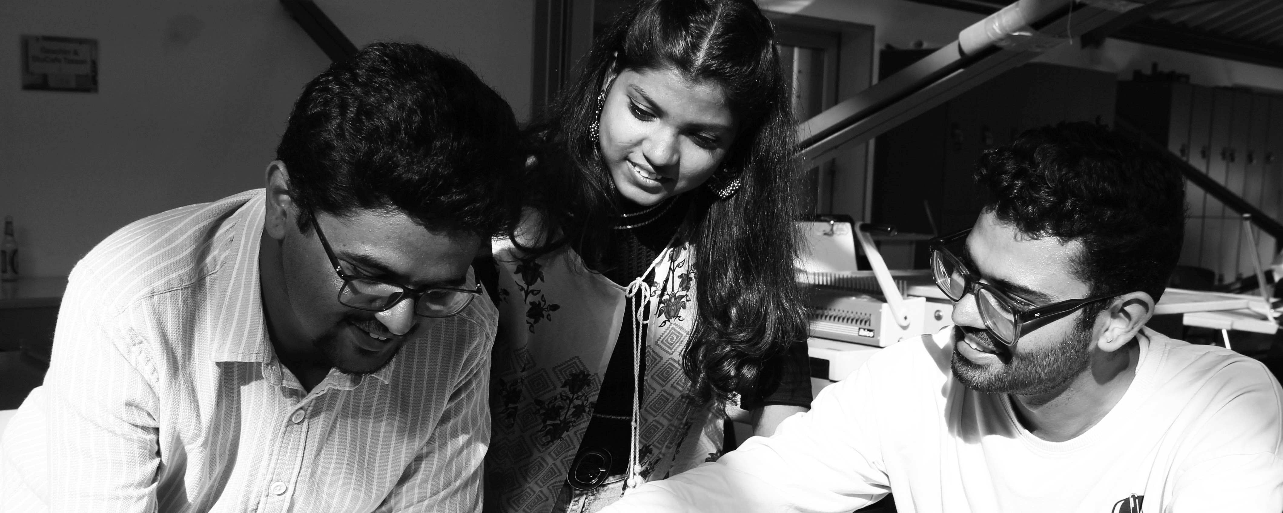 slide - Photo detail in shades of gray: Three IMLA students are leaning over a document on a table in a room. The student on the right is sitting; the other two are standing. In the background, you can see a wall, a window, technical equipment, and furniture.