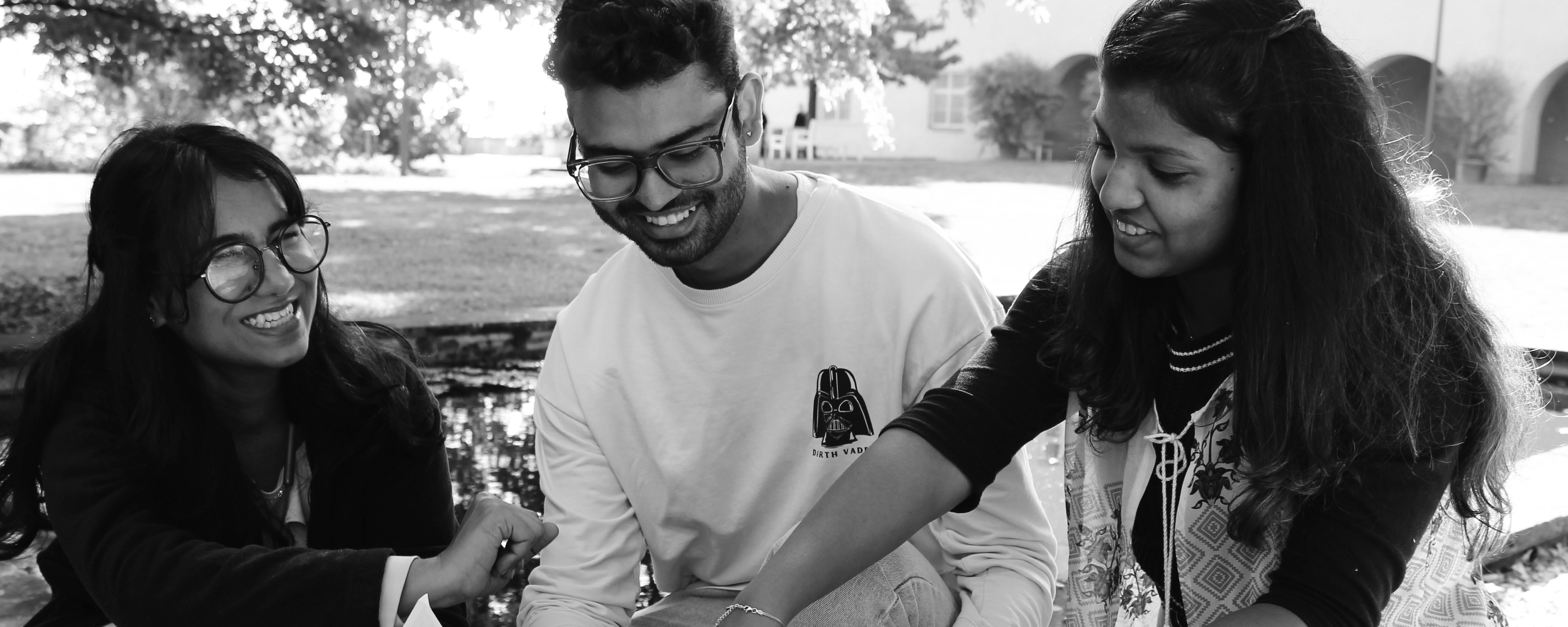 slide - Photo detail in shades of gray: Three IMLA students are sitting next to each other in a park. In the background, a body of water, a lawn, trees, and a building are visible.