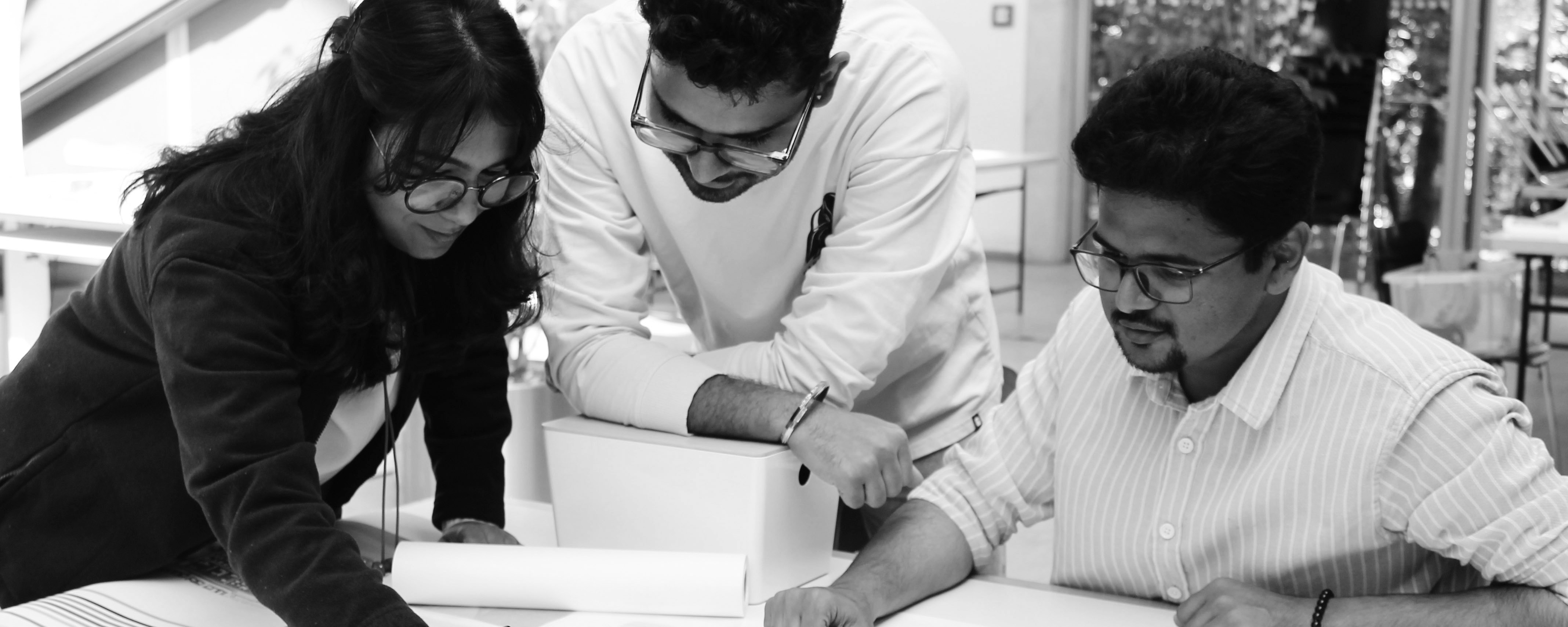 slide - Photo detail in shades of gray: Three IMLA students are leaning over a document on a table in a room. The student on the right is sitting; the other two are standing. In the background, you can see a wall, a table, and a glass door.