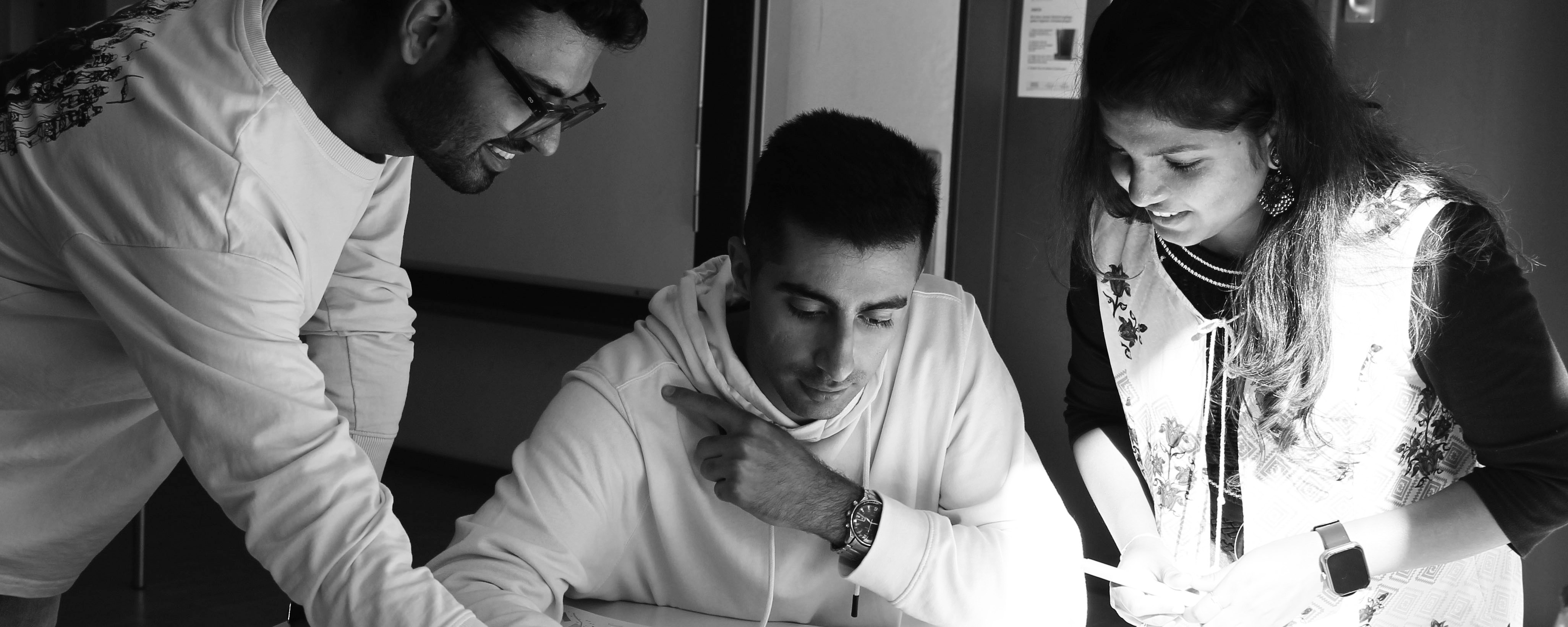 slide - Photo detail in shades of gray: Three IMLA students are leaning over a document on a table in a room. The student in the middle is sitting, while the other two are standing. A wall with a door is visible in the background.
