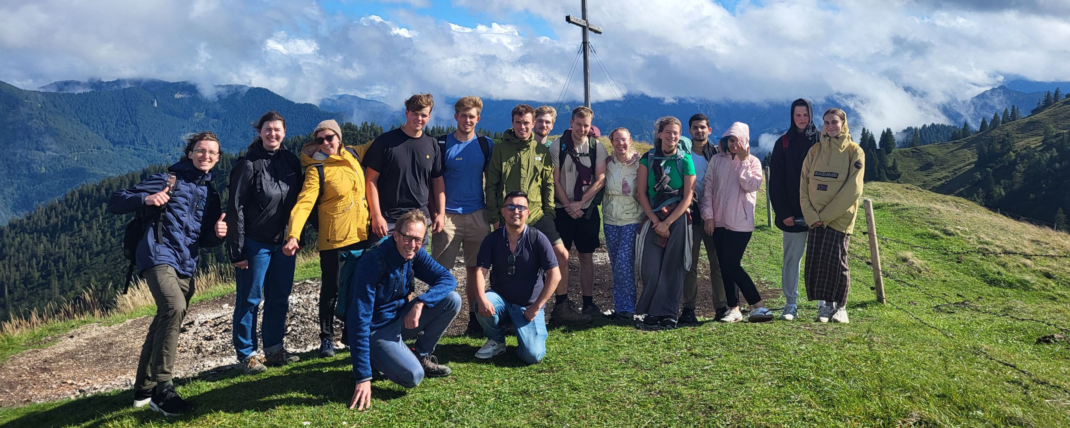 slide - Photo. Prof. Ingrid Schegk can be seen in the center of the picture, holding a certificate and a bouquet of flowers. Frederico Meireles is standing to her right. A long table and a beige wall can be seen in the background.