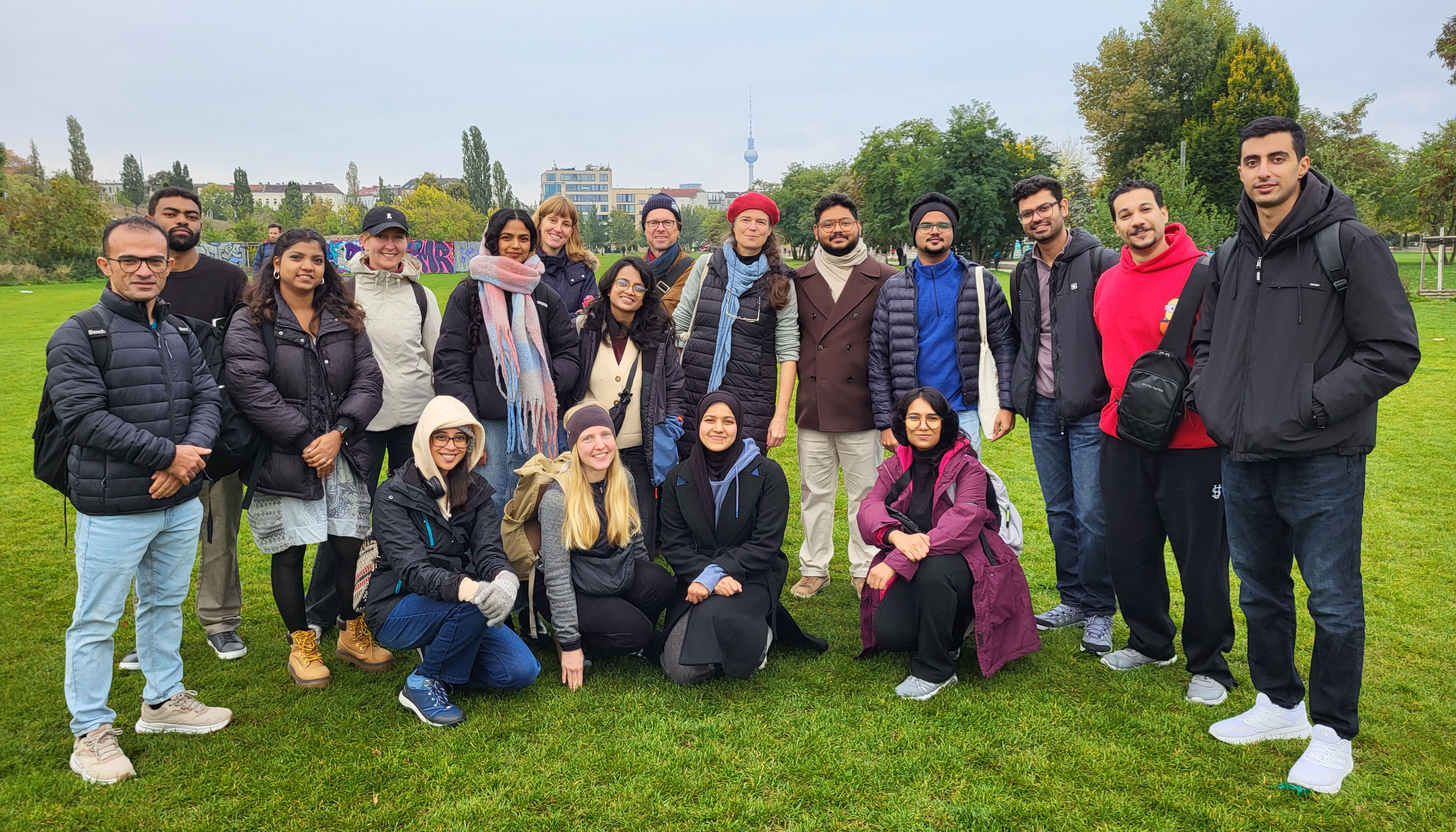 A close-up photo of a group of students and faculty members on a lawn. One row is kneeling, one row is standing, and all are facing the camera. Trees and buildings can be seen in the background