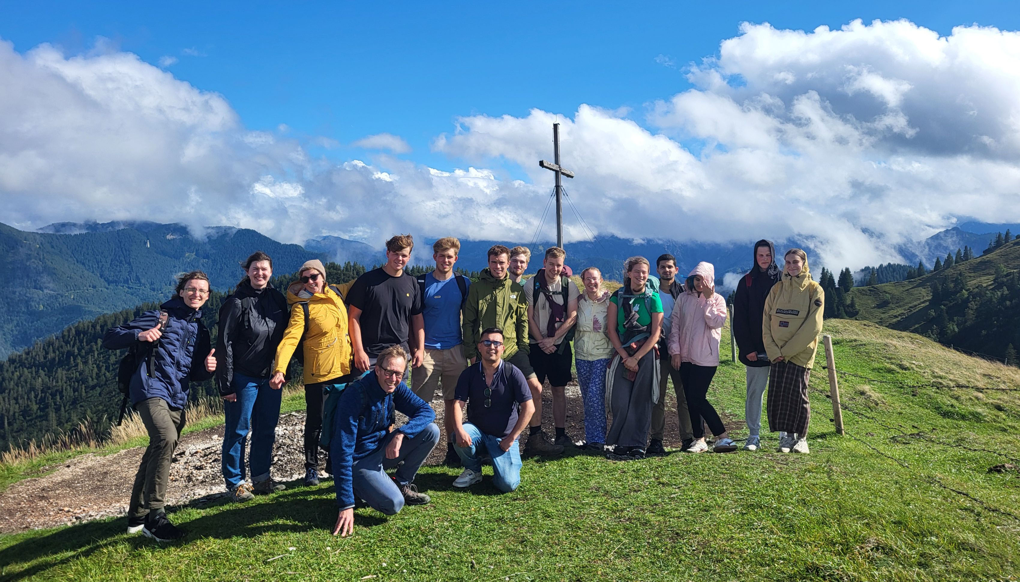 The photo shows a group of people standing in a row on a grassy summit. In the background, you can see a summit cross, forested mountains, clouds, and blue sky.