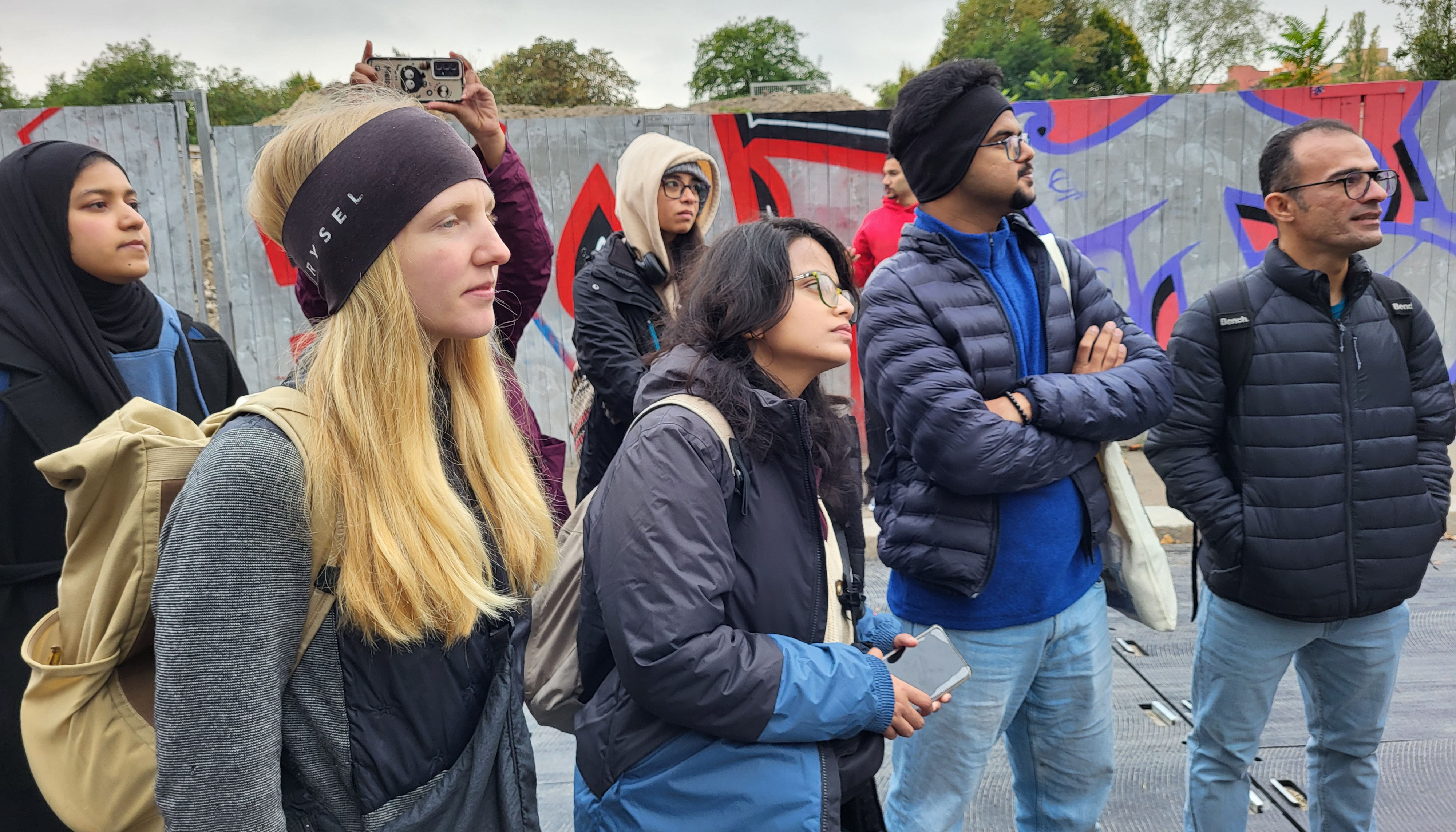 This is a cropped photo showing a group of students standing and looking to the right. A construction fence and trees can be seen in the background.