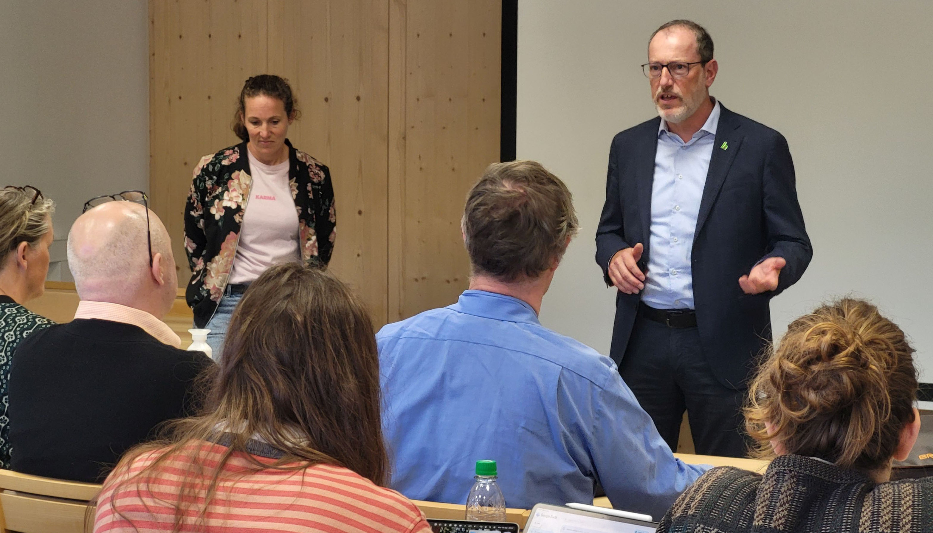 Photo detail. Audience members are seated in the foreground. Two speakers are standing in front of them. A wall can be seen in the background.