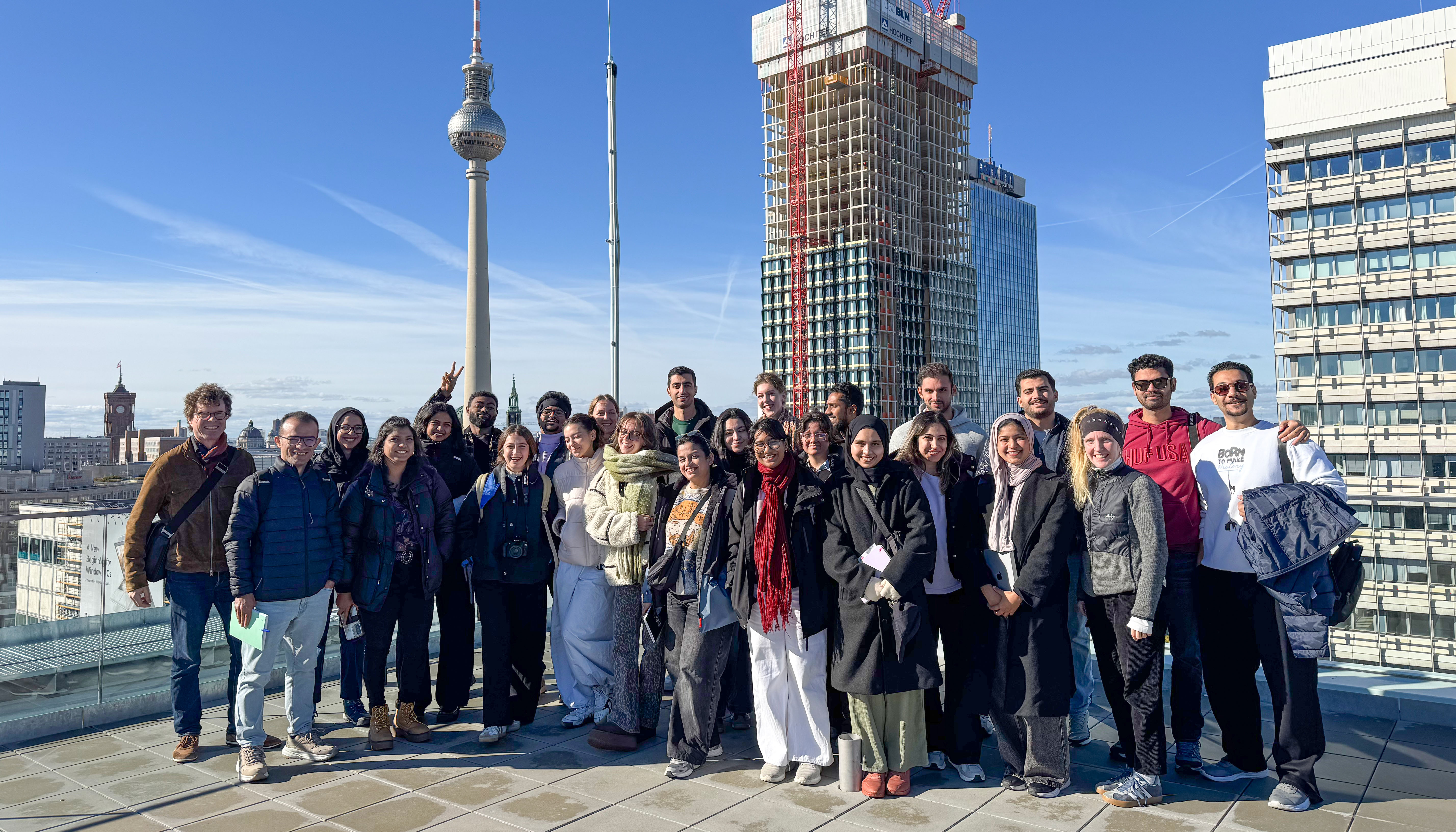 A close-up photo of a group of students and faculty members standing in two rows on a roof terrace. Everyone is facing the camera. Buildings and the sky are visible in the background.