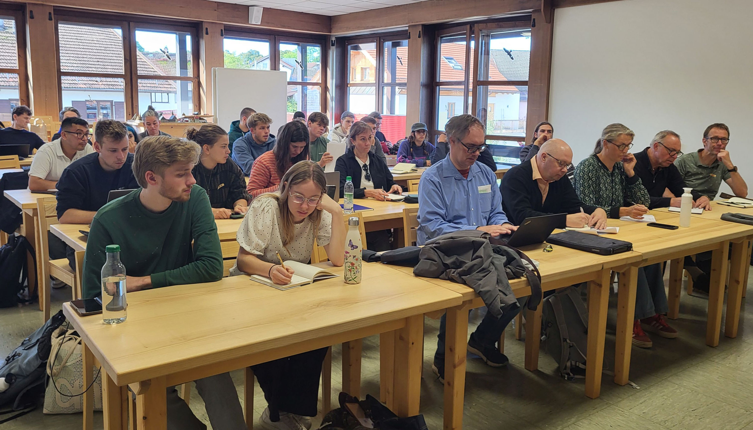The photo shows a seminar room with wooden tables and chairs where people are sitting. Windows can be seen in the background