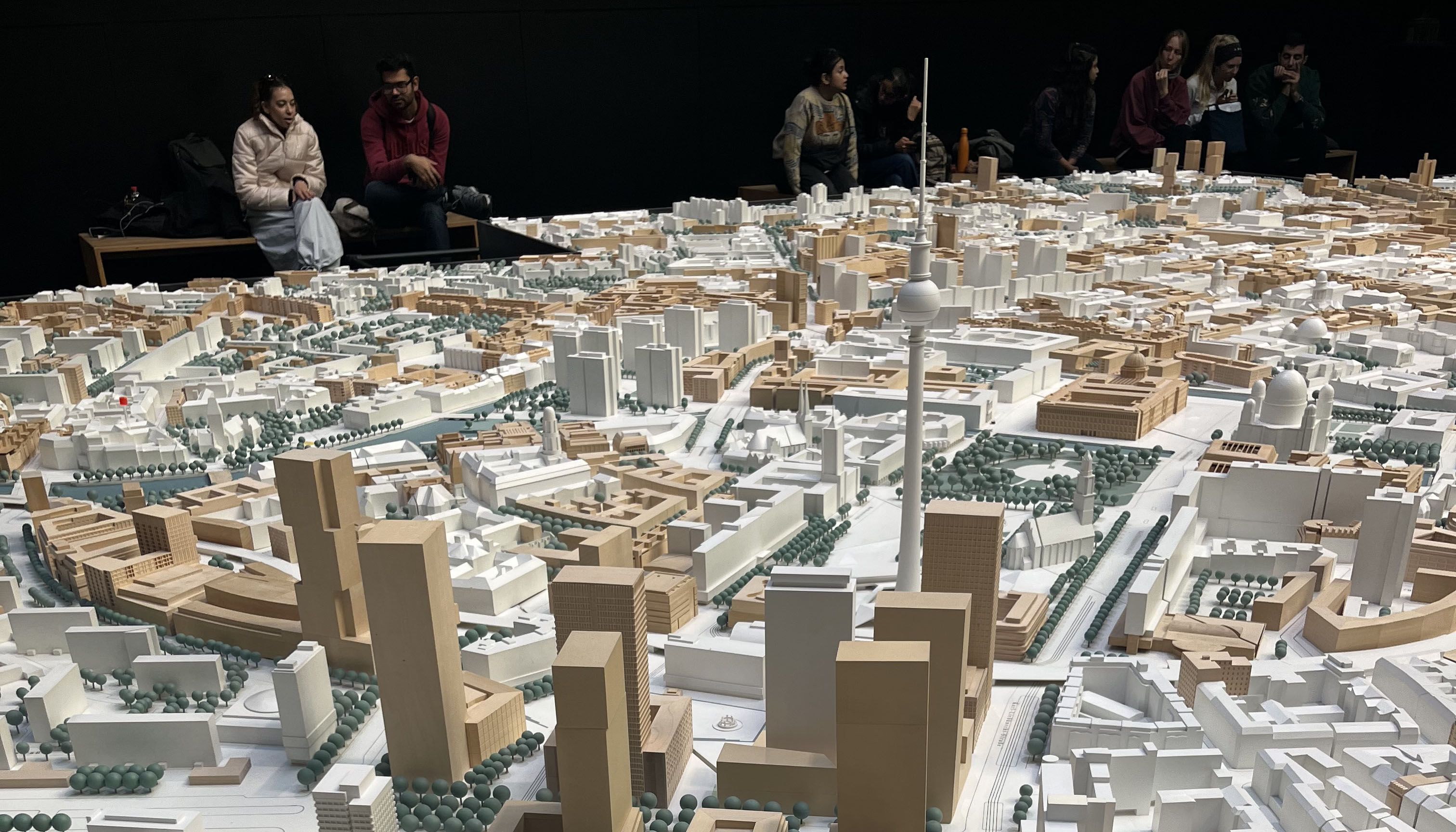 The image shows a close-up of a three-dimensional city model in the foreground. In the background, people can be seen sitting on benches.