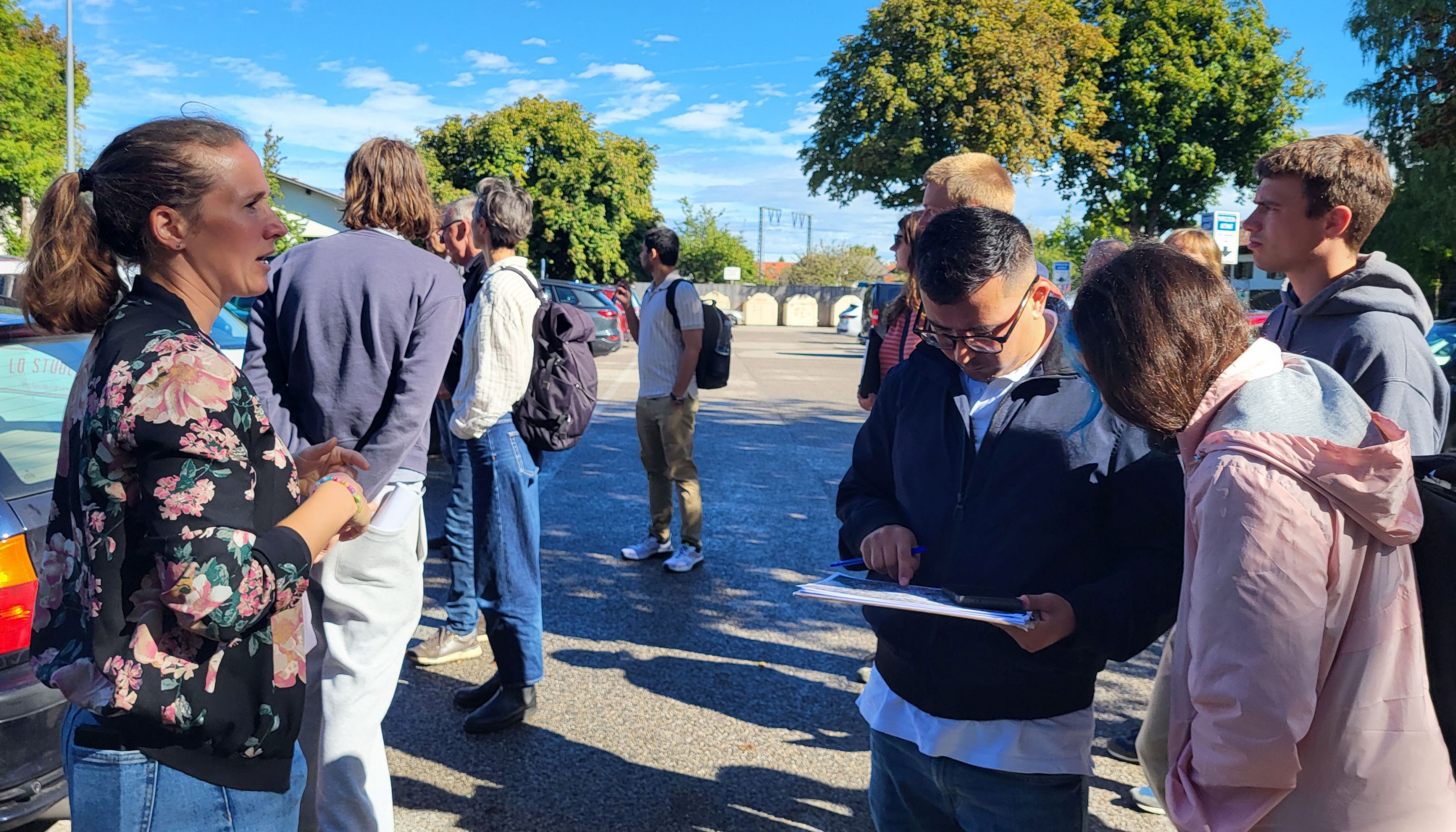 Photo detail of a group of people in a parking lot. Trees and buildings can be seen in the background.