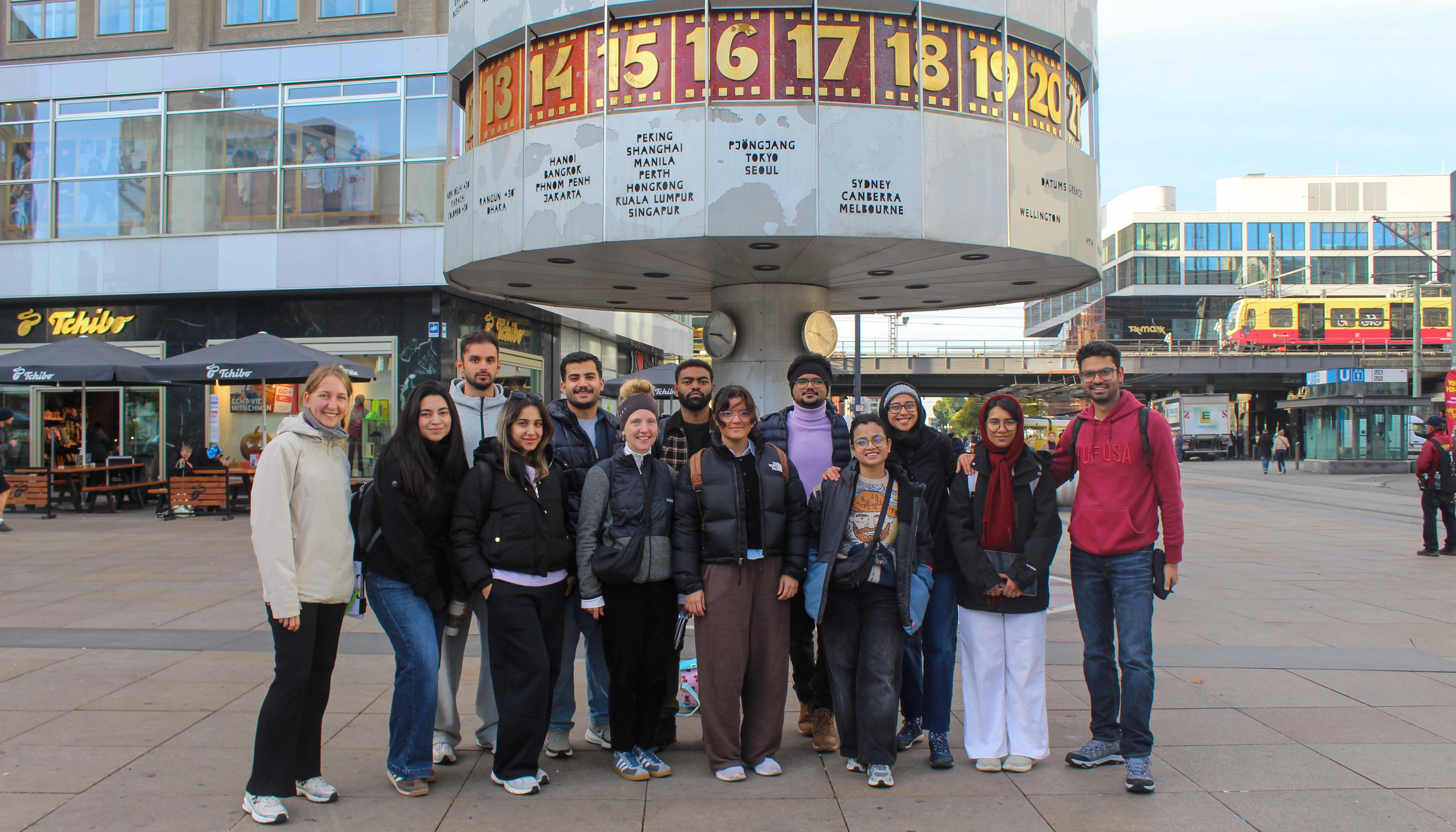 The photo shows a close-up of a group of students standing in a row on a concrete-paved plaza in the foreground. They are all facing the camera. A world clock and buildings can be seen in the background.