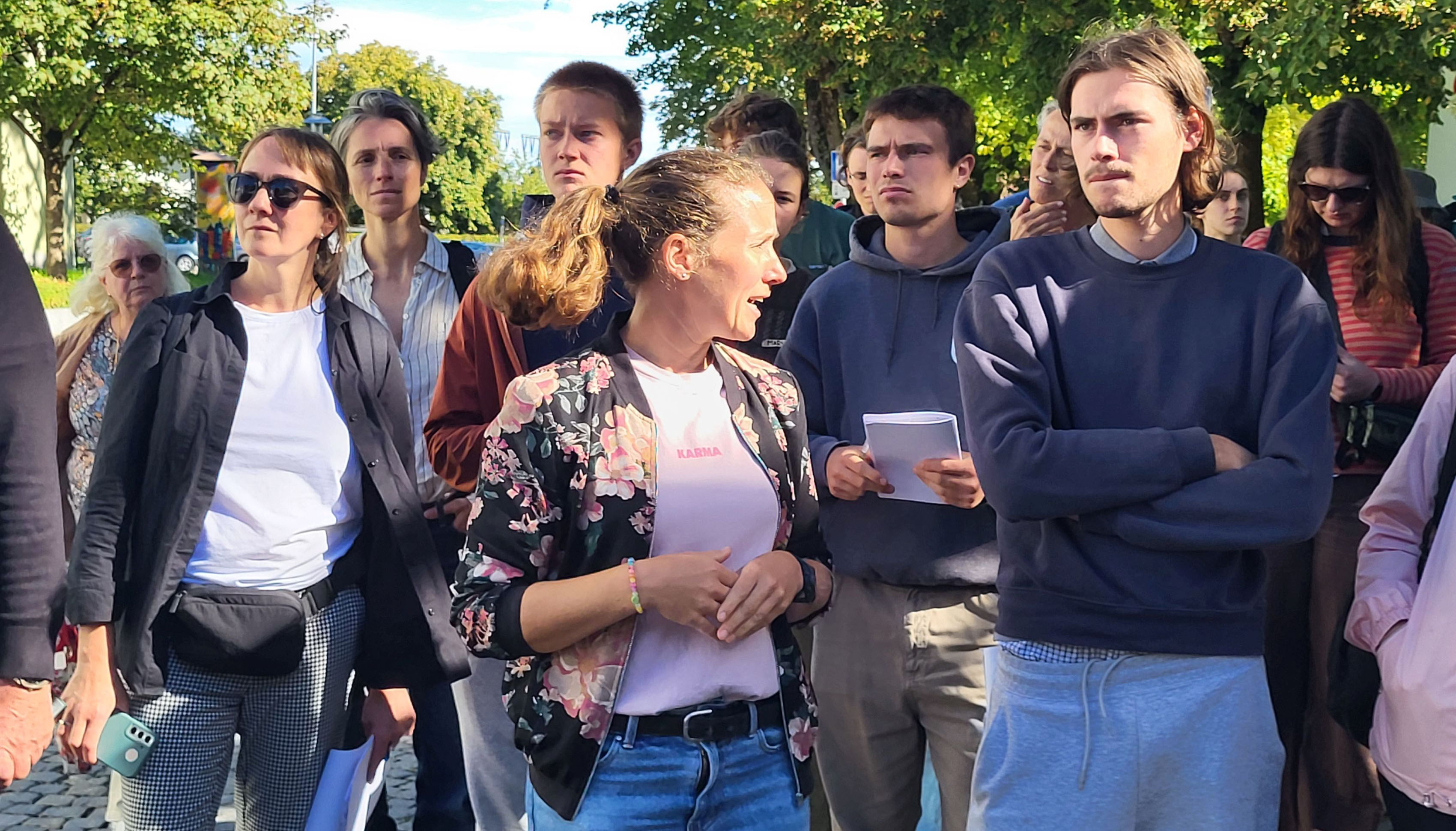 Photo detail of a group of people standing in a row, looking in one direction and photographed from the front. Trees can be seen in the background.
