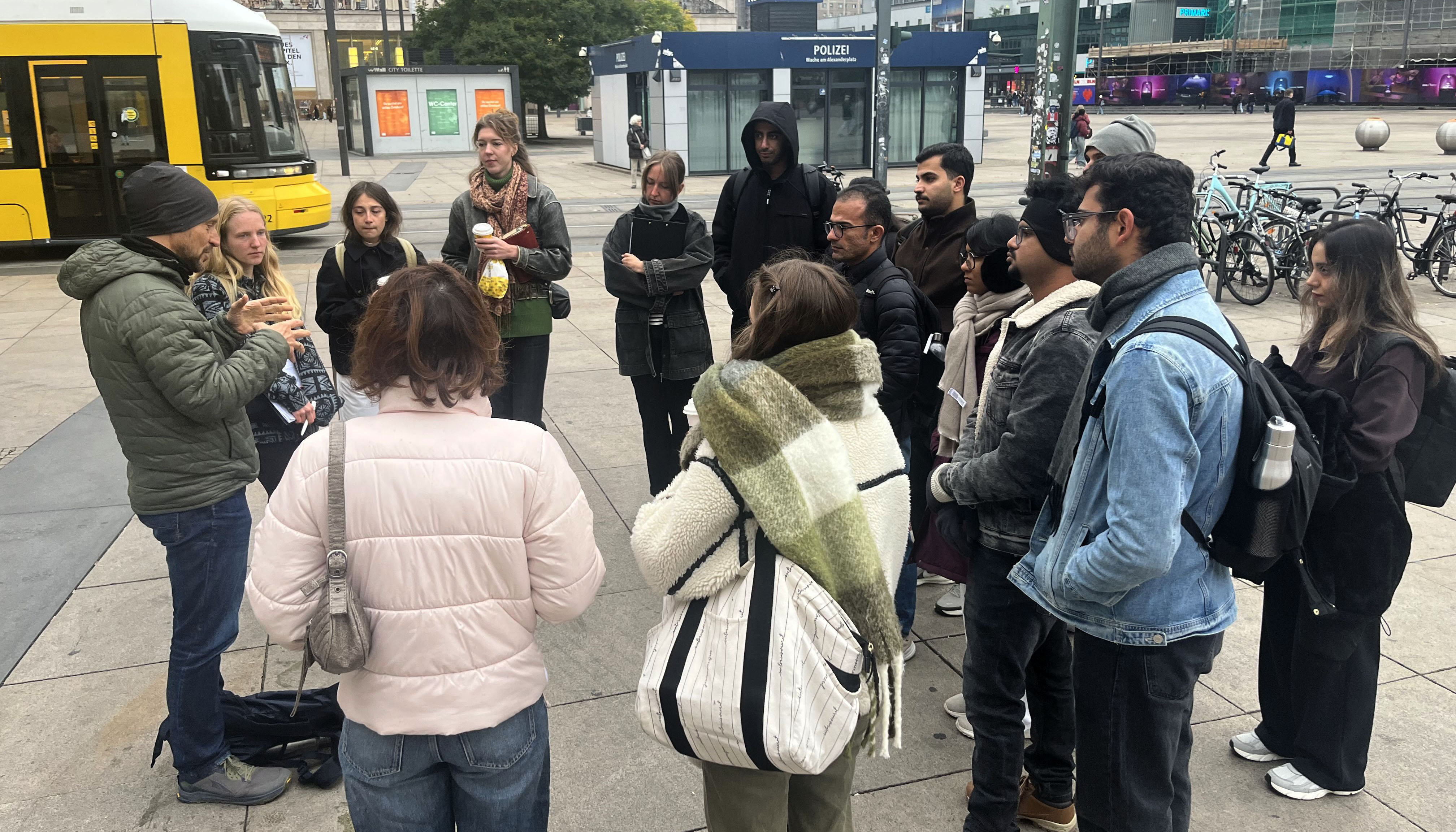 The photo shows a group of students standing in a circle on a concrete-paved plaza, listening to a speaker. A streetcar and buildings can be seen in the background.
