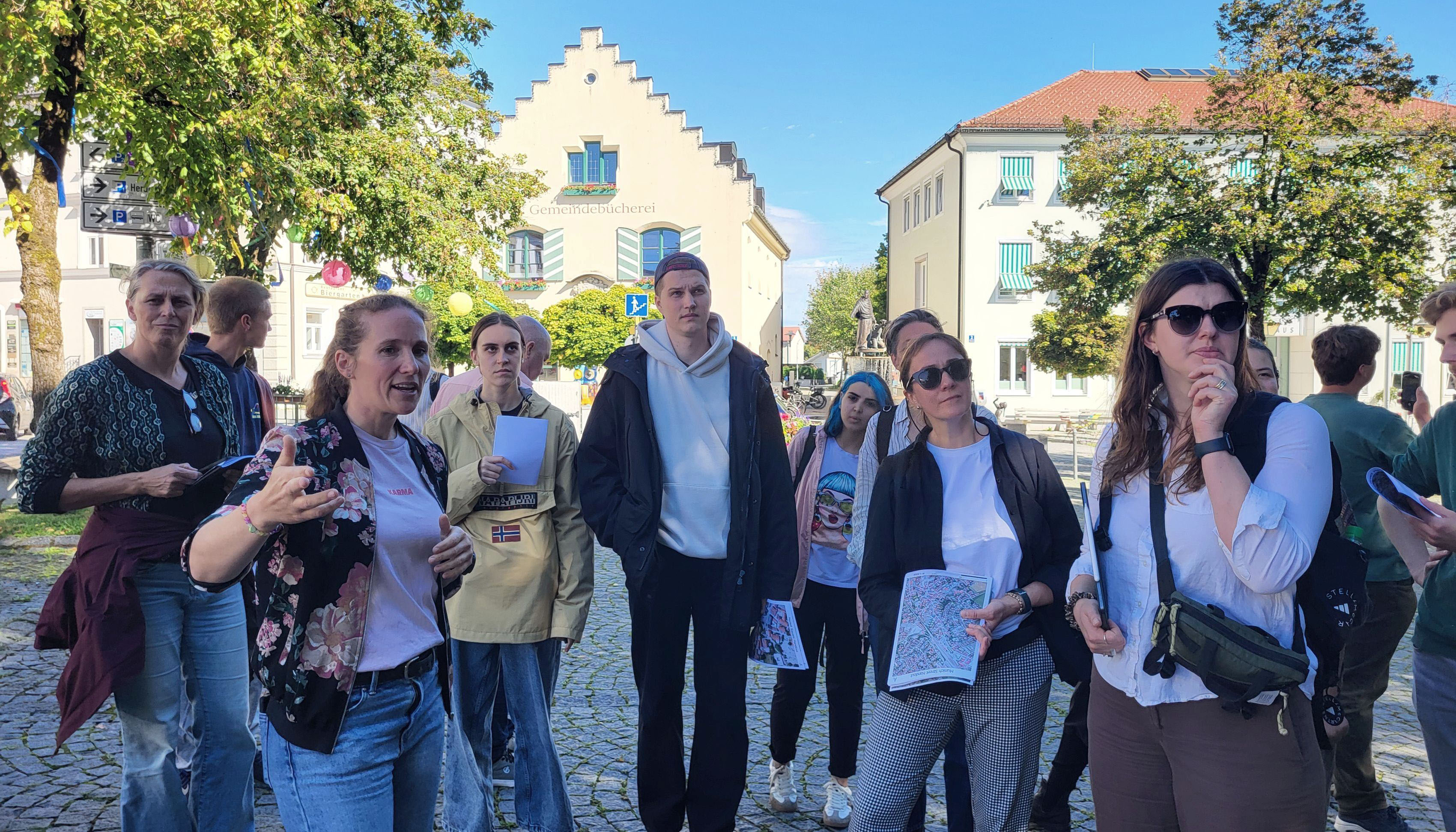 Photo detail of a group of people standing in a row, looking in one direction and photographed from the front. Trees and buildings can be seen in the background.