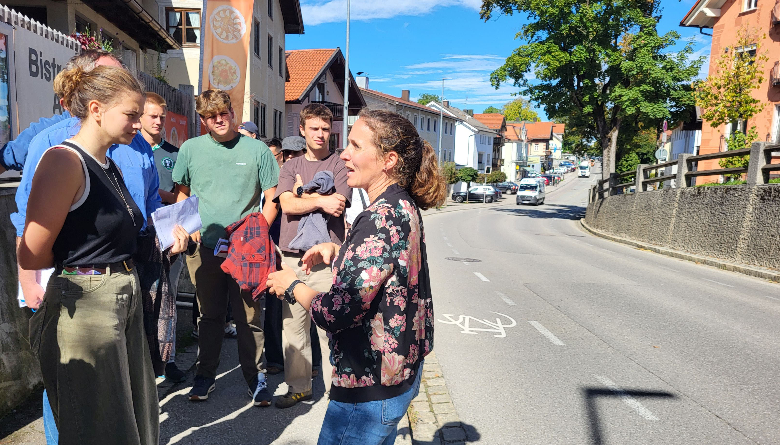 The photo shows a group of people standing on a sidewalk. There is a road on the right-hand side, while buildings and a tree can be seen in the background.