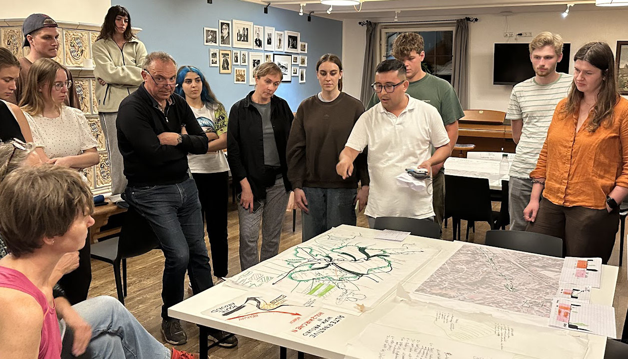 The photo shows a group of people standing around a table on which plans are laid out. Walls and a window can be seen in the background.