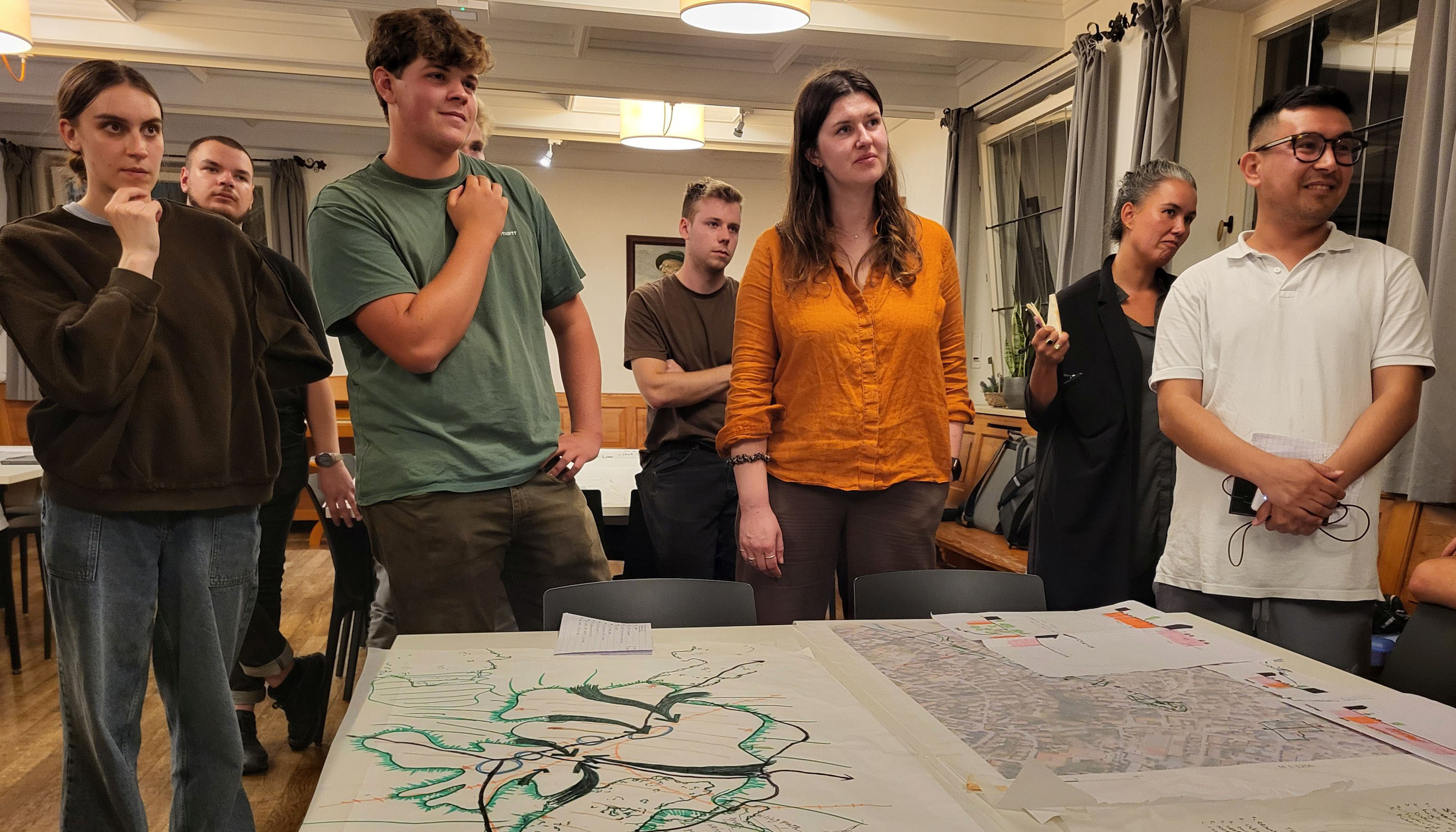 The photo shows a group of people standing behind a table on which plans are laid out. Walls and windows can be seen in the background.