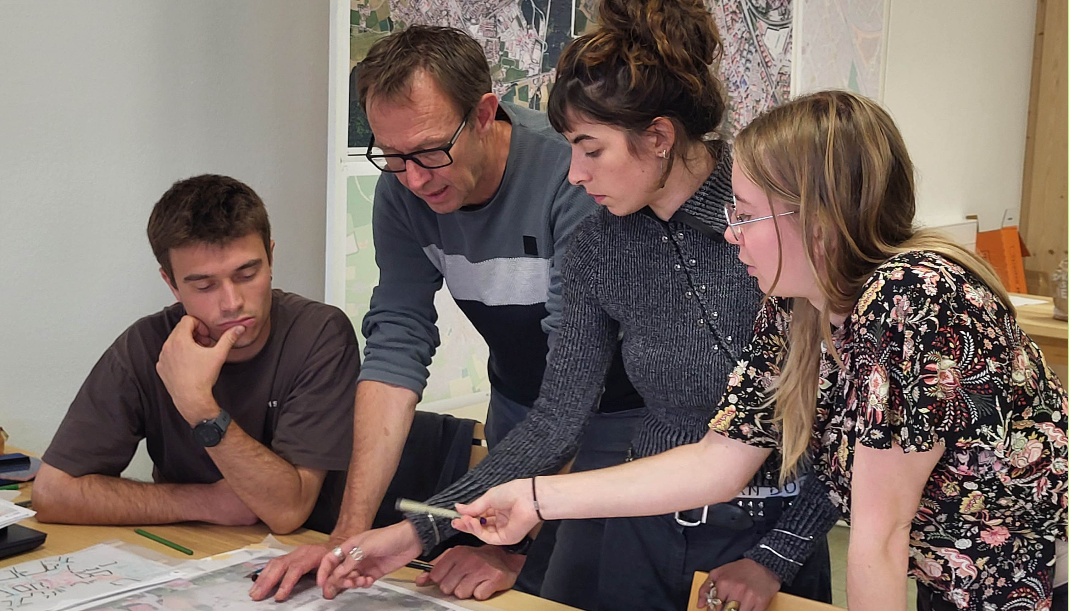 The photo shows four people leaning over a table covered with plans. In the background, you can see a wall with plans pinned to it.