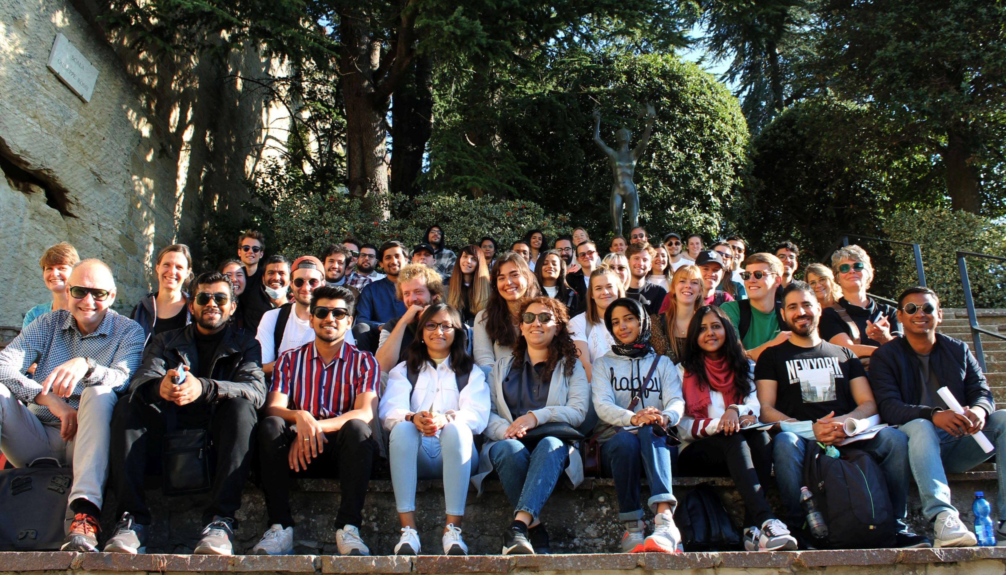 Photo group of students and lecturers, sitting on stairs, background statue and trees