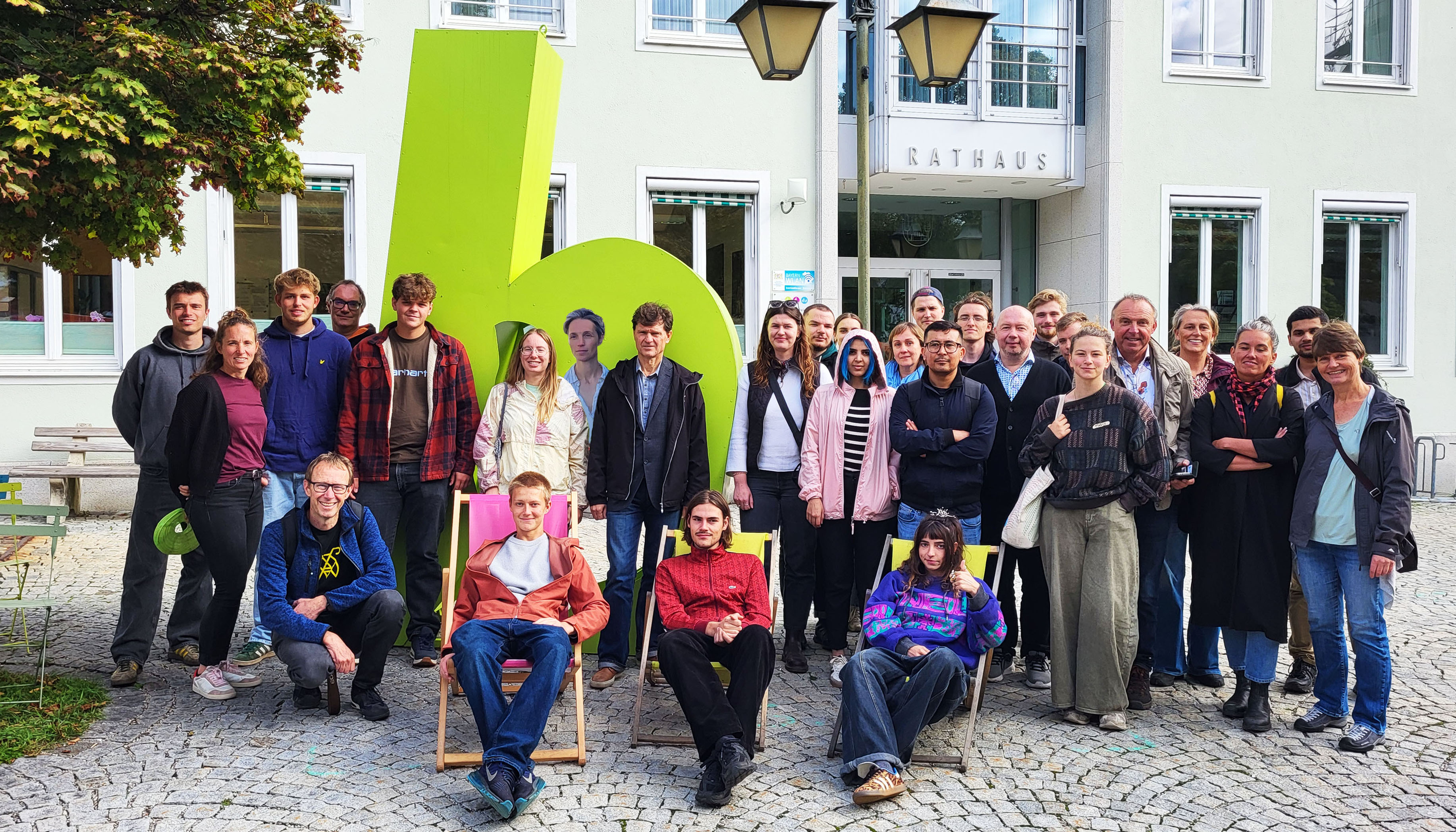 The group photo shows a square paved with gray natural stone. Three people are sitting in deck chairs in the front row. Behind them is another row of people. In the background, there is a green sculpture representing a "b", a lantern, and a building with the inscription “Rathaus” (town hall).