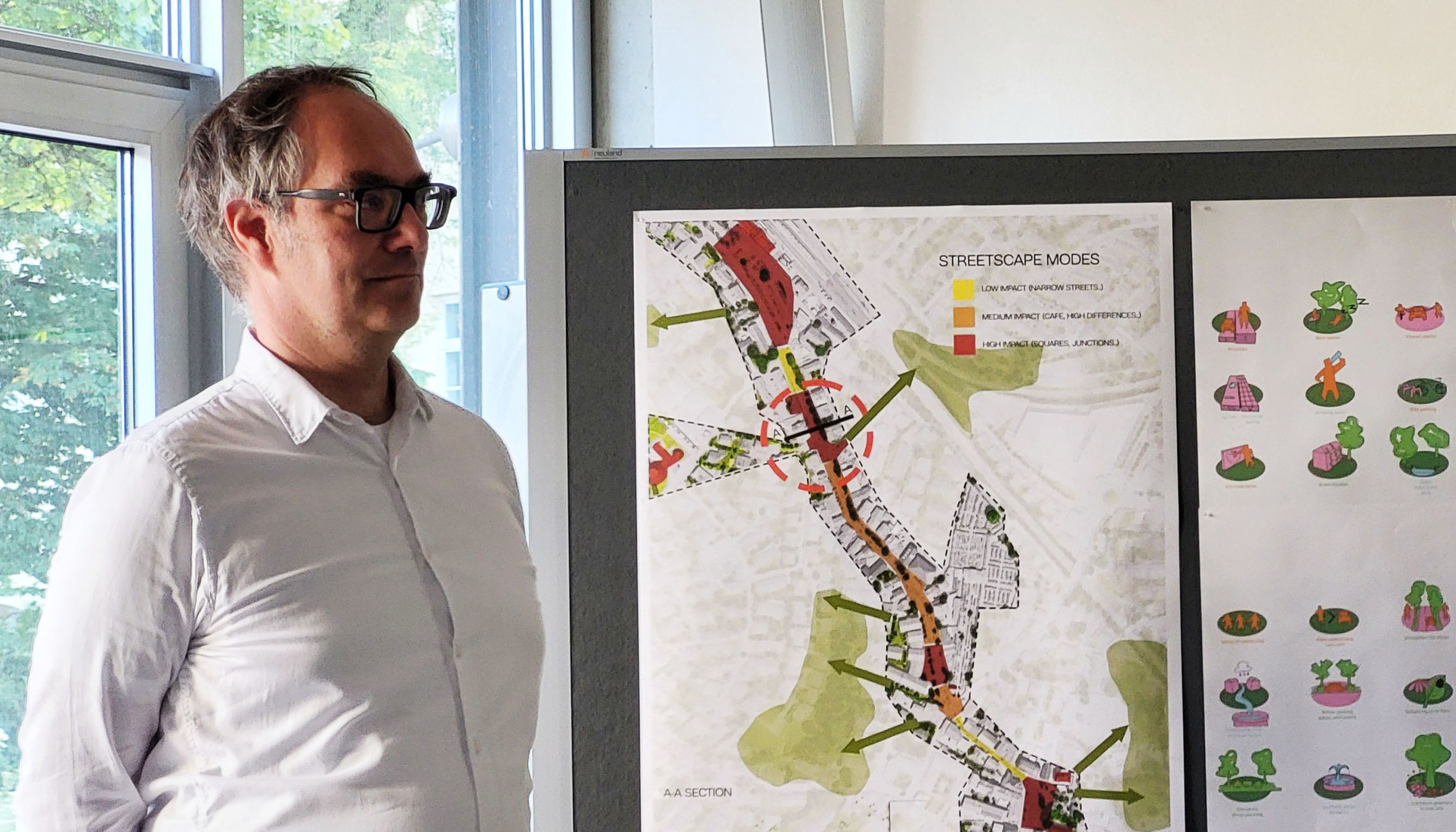 Photo detail. On the left, a person can be seen standing in front of a window. To the right of them is a display board with plans.