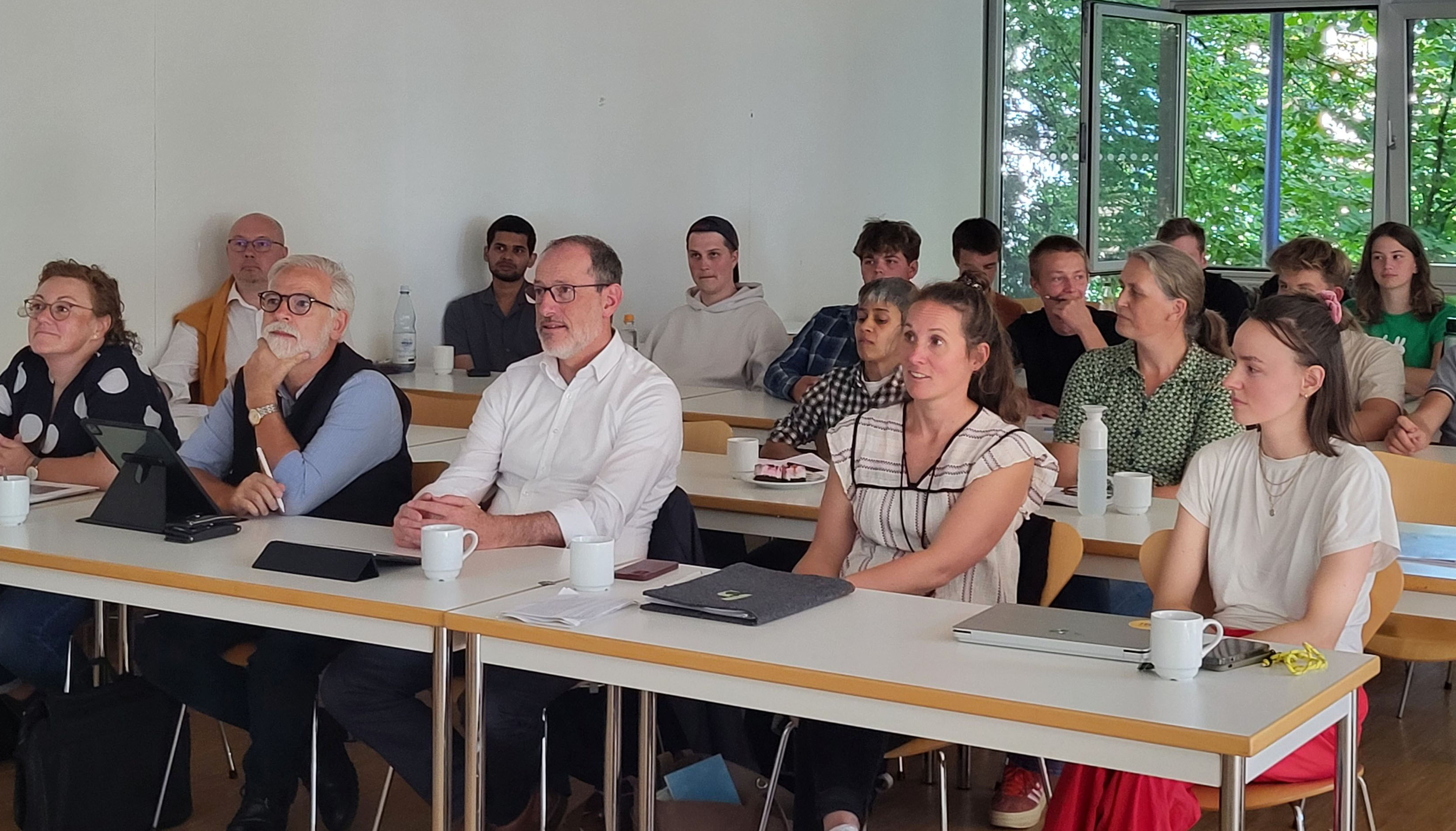 The photo shows a seminar room with tables and chairs where people are sitting. A wall and a window can be seen in the background.