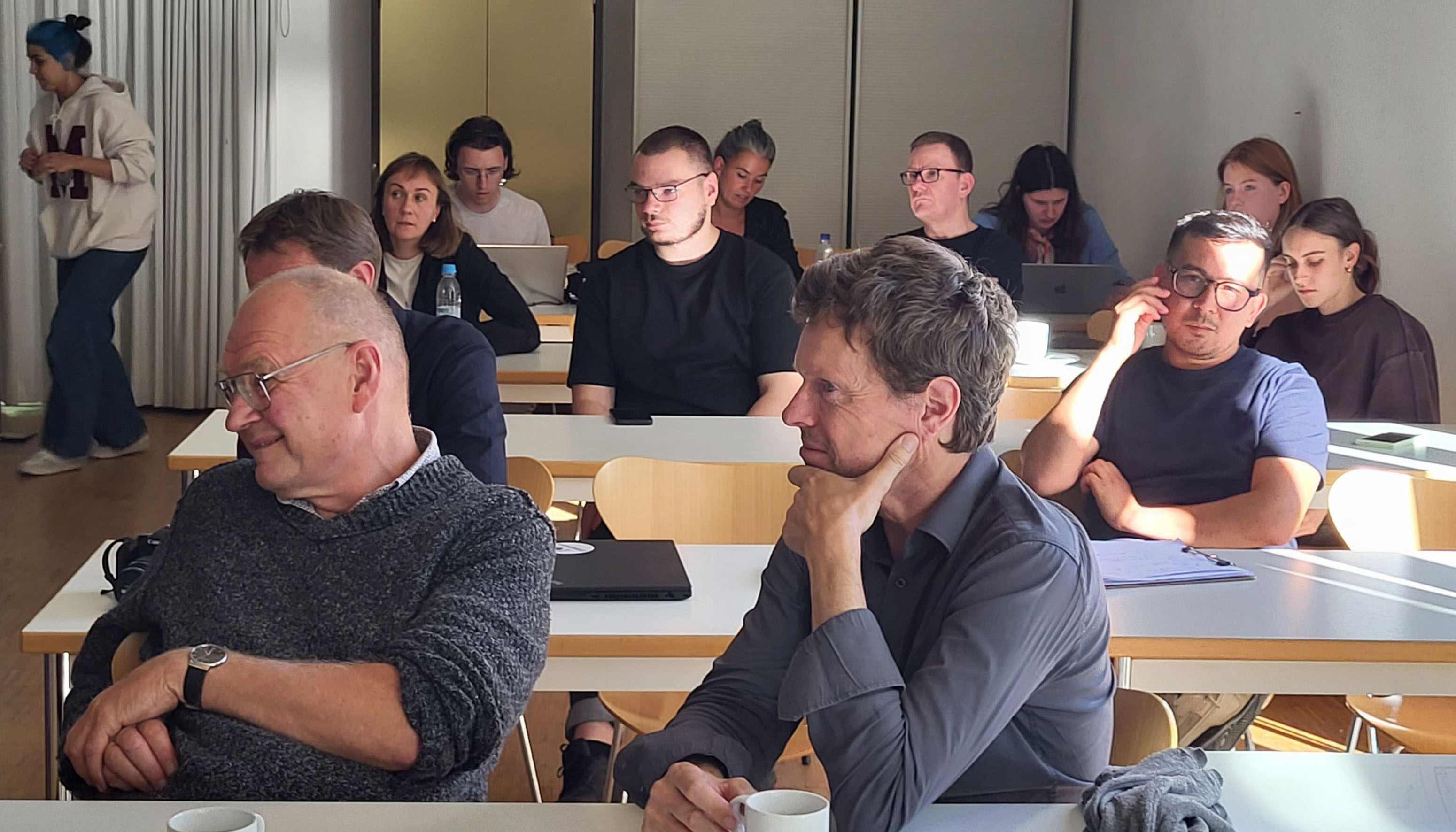 The photo shows a seminar room with tables and chairs where people are sitting. A wall and a cupboard can be seen in the background.