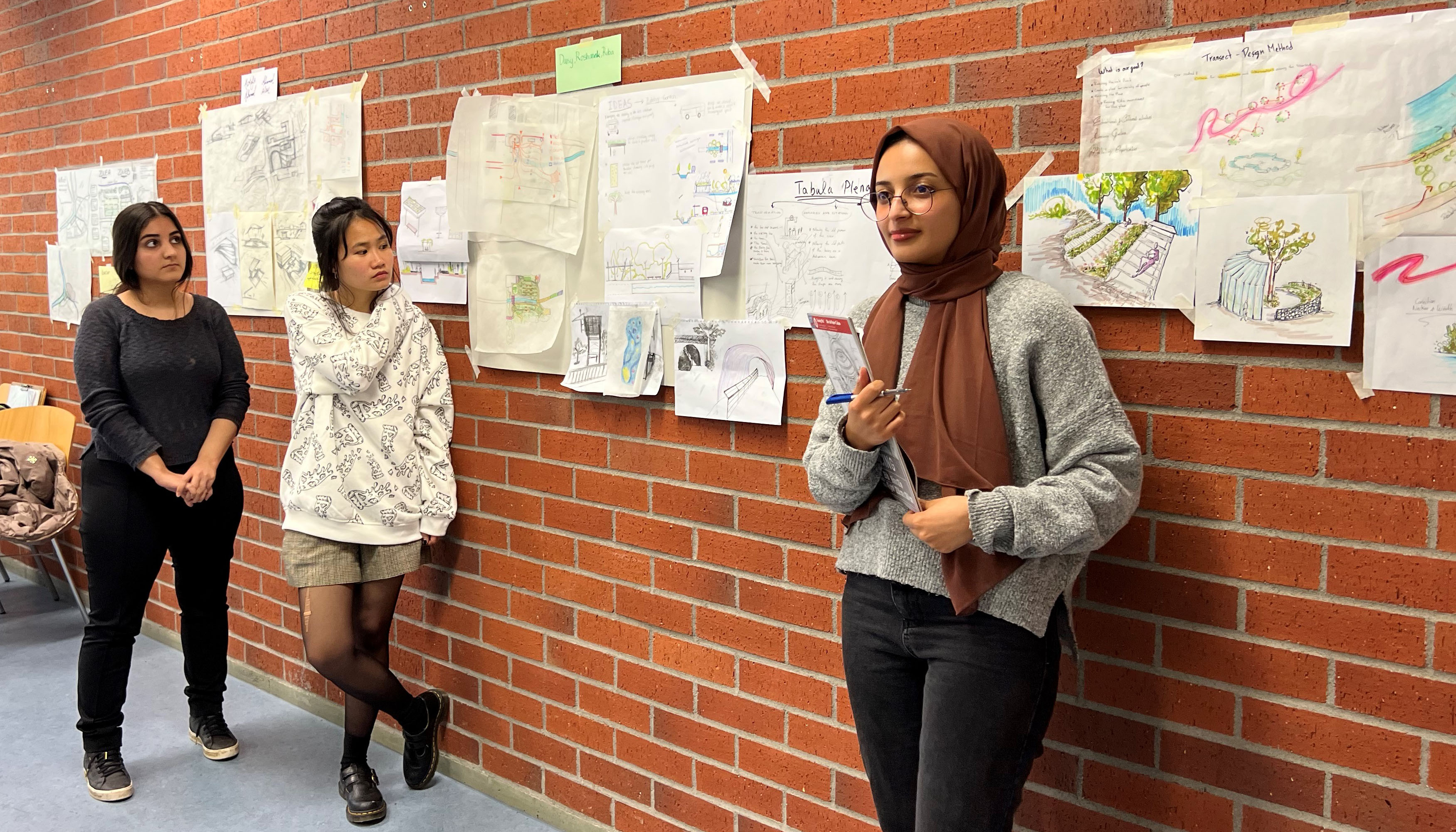 Photo detail of three female students in front of a wall with sketches and plans.