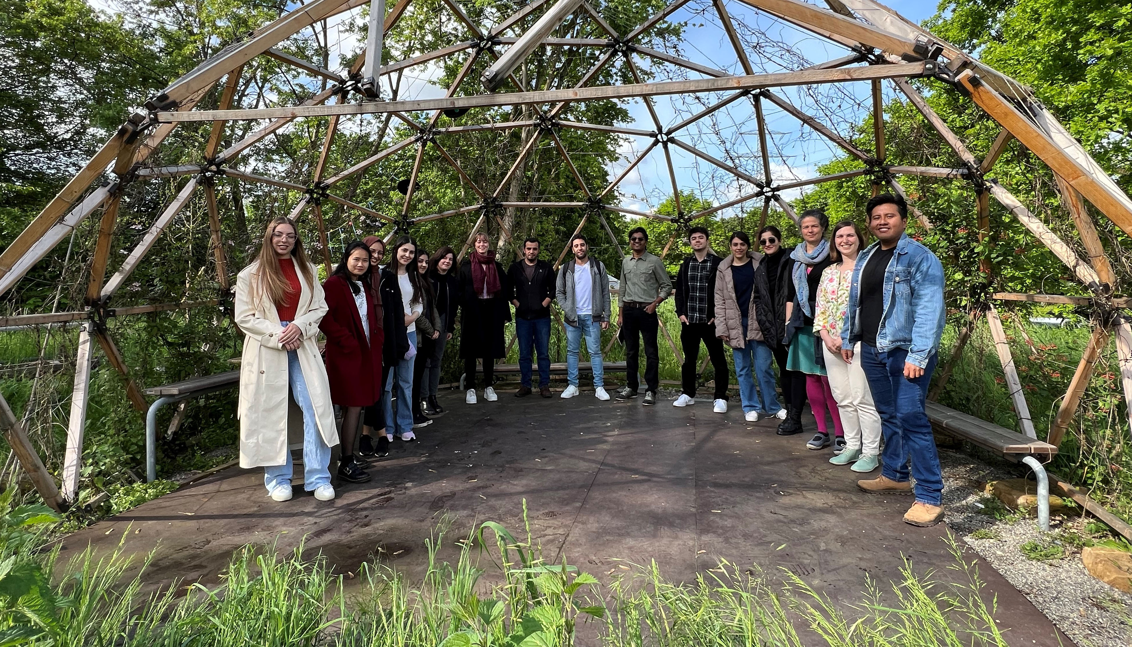Group of students around a slabbed square, square on a meadow, covered by a wooden dome, trees in the background.