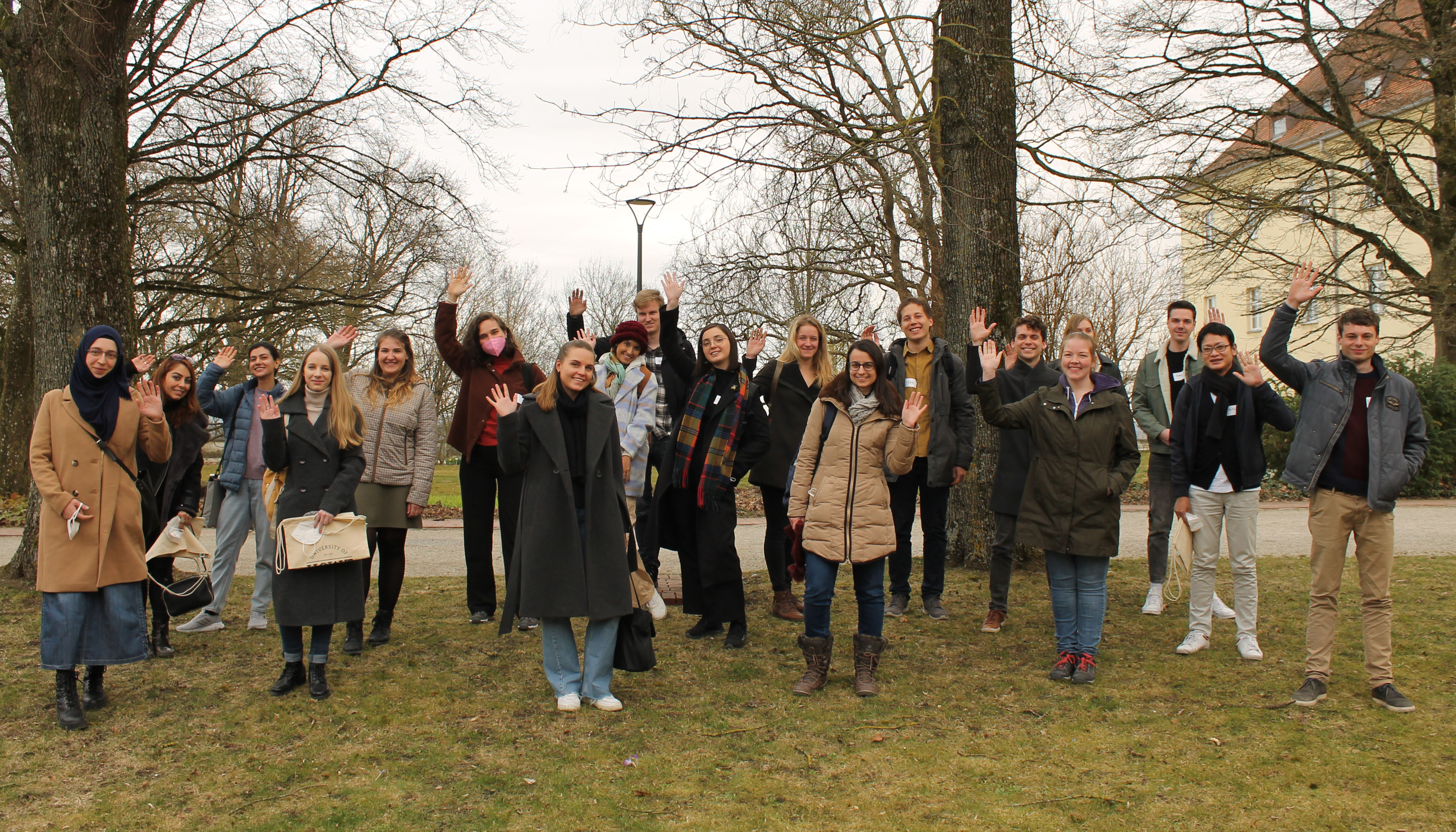 Photo group of students, waving, on lawn, background trees and bushes