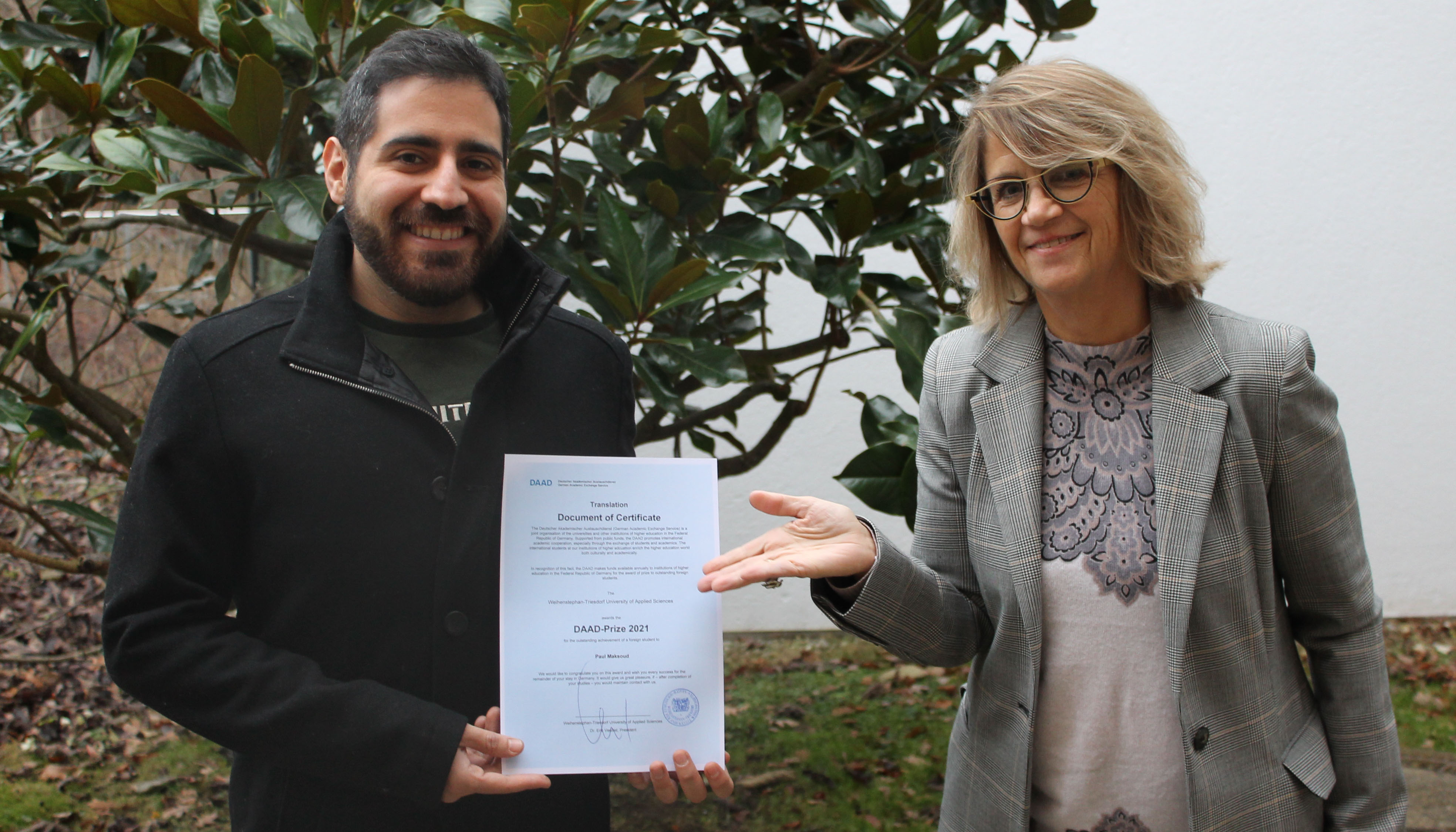 Photo in the garden, background shrub. Paul Maksoud with certificate (left in picture), Prof. Ingrid Schegk (right in picture).